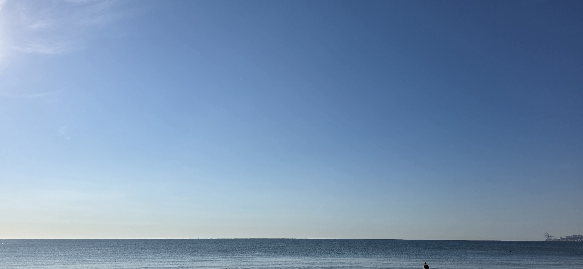 The image depicts a serene seascape with a vast expanse of clear blue sky dominating the upper portion. The horizon line is sharp, dividing the sky from the calm ocean. The water is a deep blue, with subtle ripples indicating a gentle breeze. In the distance, along the horizon, a faint silhouette of industrial structures, possibly cranes at a port, can be observed on the right side. A solitary figure is visible in the water near the bottom center of the frame, appearing to be wading or swimming. The overall impression is one of tranquility and open space.