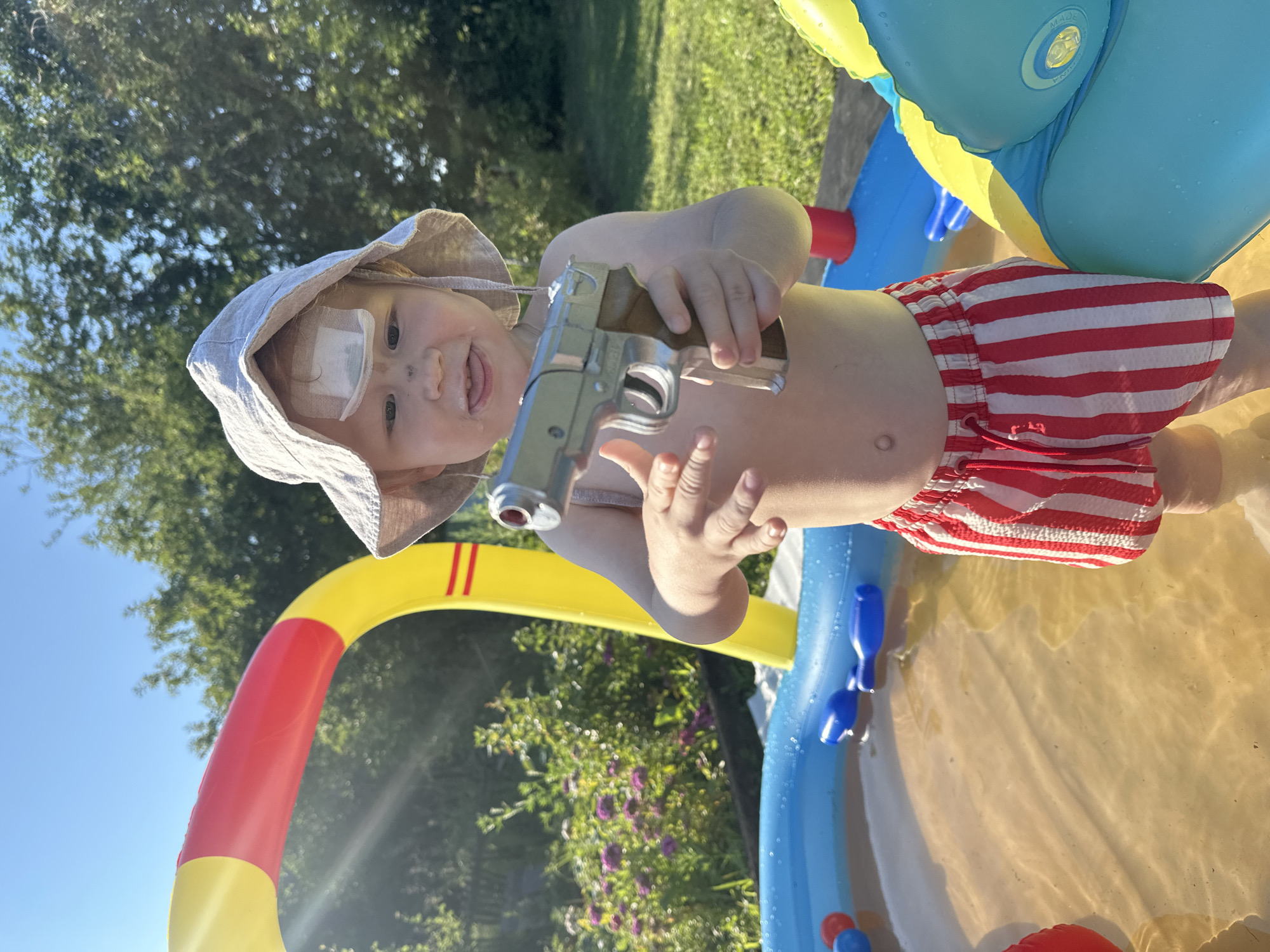 The image depicts a young child standing in an inflatable kiddie pool in a grassy backyard. The child is wearing a light-colored sun hat and red and white striped swim trunks. The child has a bandage on their forehead and smudges of dirt on their face. They are holding a silver toy water gun. The pool contains water and several blue toy objects. In the background, there is lush green foliage and trees, suggesting a sunny day outdoors. The specific location within the city of Galevo, Russia, cannot be determined from the image alone.