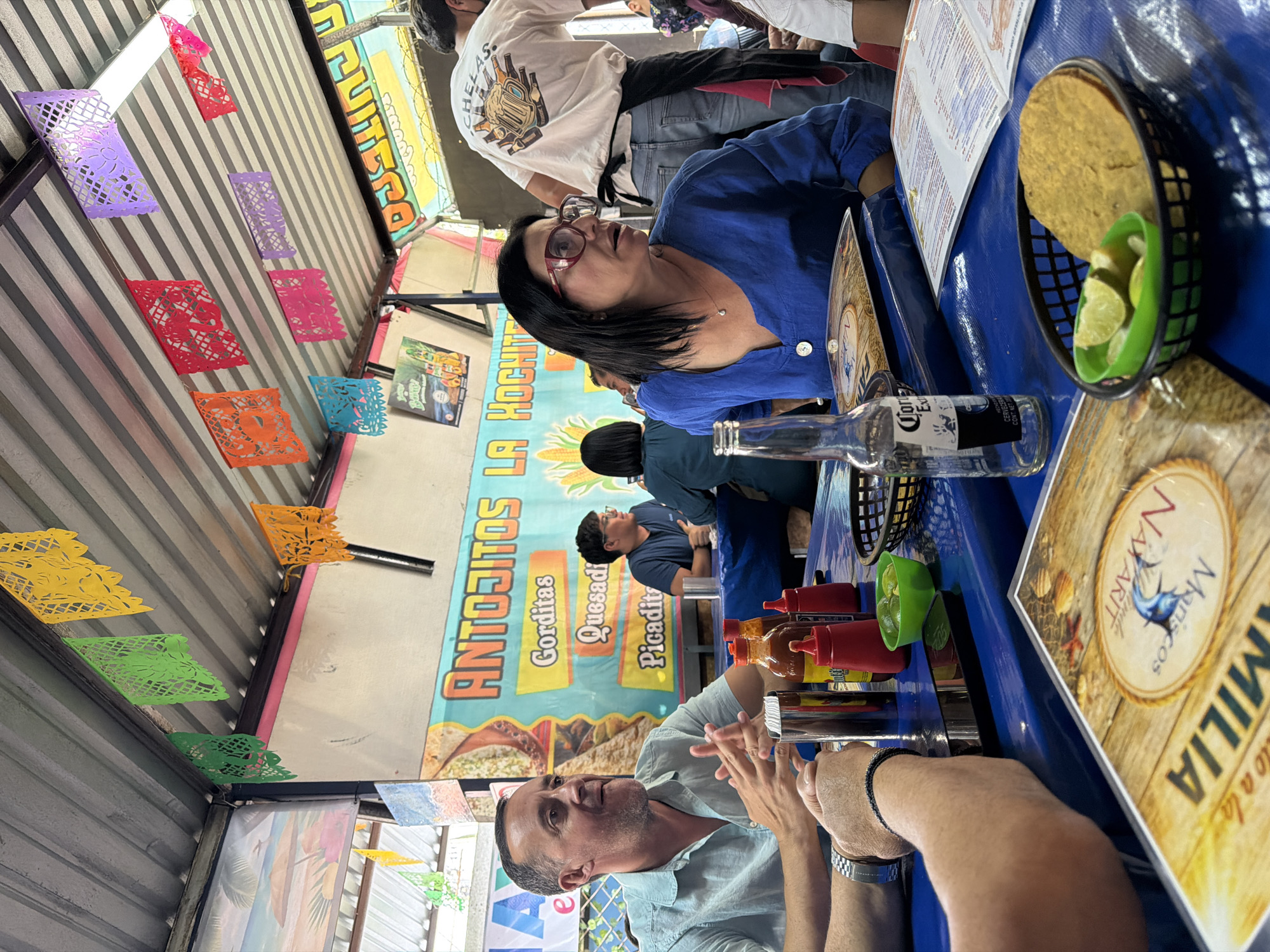 The image shows an outdoor dining scene, likely in San Andrés Cholula, Mexico. The setting appears to be a casual restaurant or food stall, as suggested by the colorful papel picado decorations hanging from the ceiling, and a banner advertising "Antojitos La Hochi" with various Mexican snacks like Gorditas, Quesadillas, and Picaditas. People are seated at a blue table. A woman with glasses wearing a blue shirt is talking to other members of her party. A man with a light blue shirt is seated at the table, as well as other people are partially visible in the background. On the table, there is a bottle of Corona Extra beer, a basket with tortillas and limes, bottles of hot sauce, and a menu with the restaurant's name "Familia Nayarit Mariscos" visible on it. The scene suggests a social gathering at a local establishment.