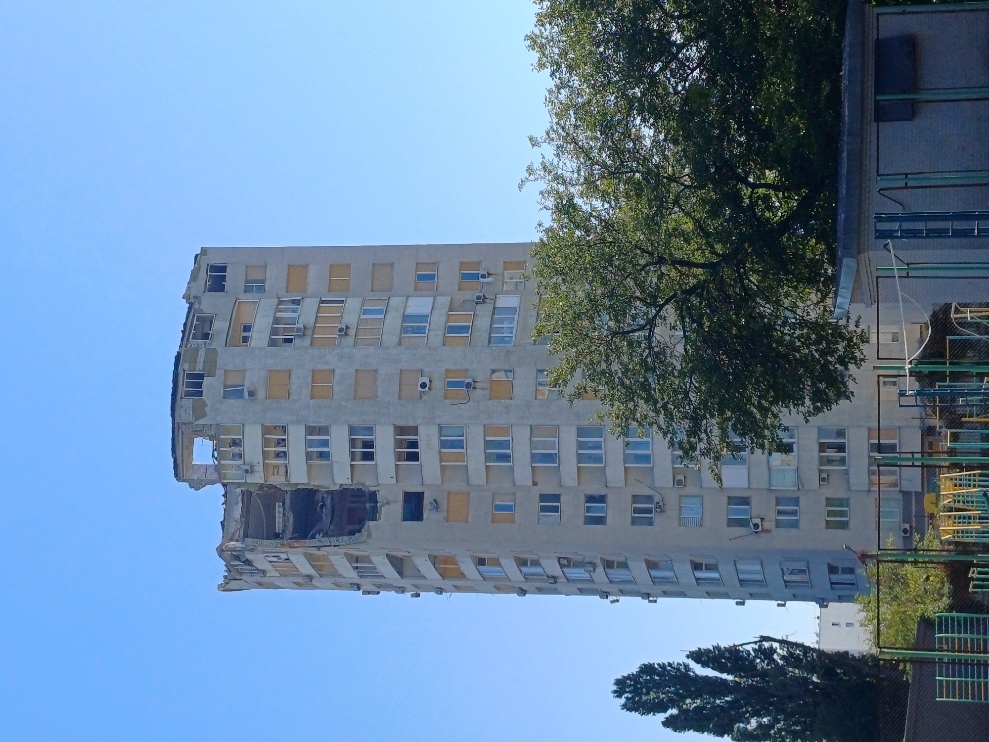 In Kyiv, Ukraine, a severely damaged high-rise apartment building stands under a clear blue sky. The top floors of the building are heavily compromised, with sections of the facade ripped away, exposing the internal structure. Many windows are broken, while others are boarded up with yellow panels. Air conditioning units are visible attached to the building's exterior. In the foreground, lush green trees partially obscure the lower portion of the building, and behind them, a low-rise structure and what appears to be a fenced-off recreational area with playground equipment are visible. The scene depicts destruction and the aftermath of an event affecting the urban environment.