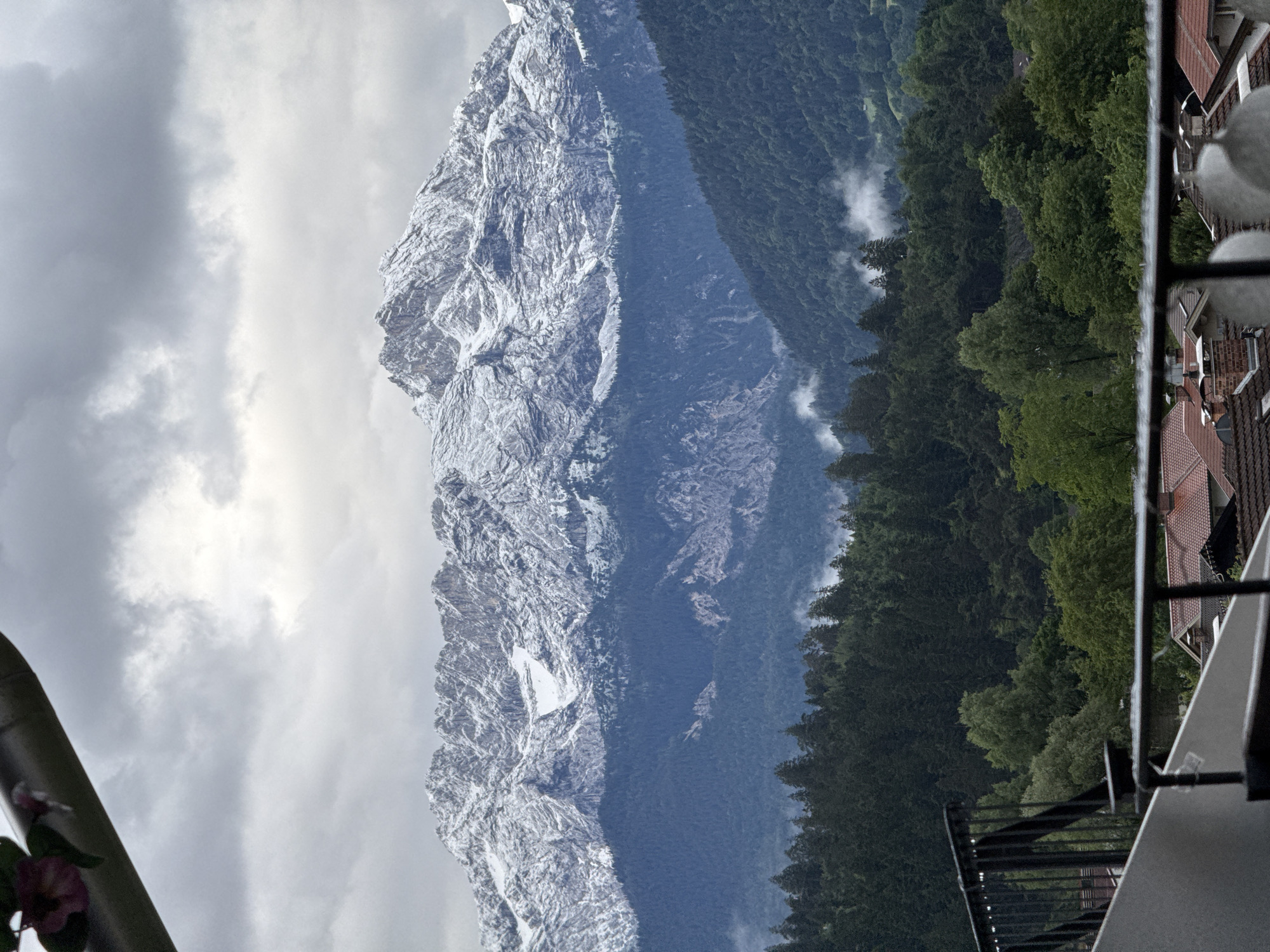 The image depicts a panoramic view of a mountainous landscape with snow-capped peaks under a cloudy sky. In the foreground, a balcony railing is visible, suggesting the vantage point is from a building in the city of Garmisch-Partenkirchen, Germany. The mountains are covered in a mixture of snow and rock, with dense evergreen forests dominating the lower slopes. Patches of fog are present in the valleys, adding to the atmospheric quality of the scene. Below the forested mountains, the rooftops of houses are visible, indicating the presence of a town or village. No people are discernible in the image. The overall impression is one of a serene, natural environment with human habitation nestled at its base.