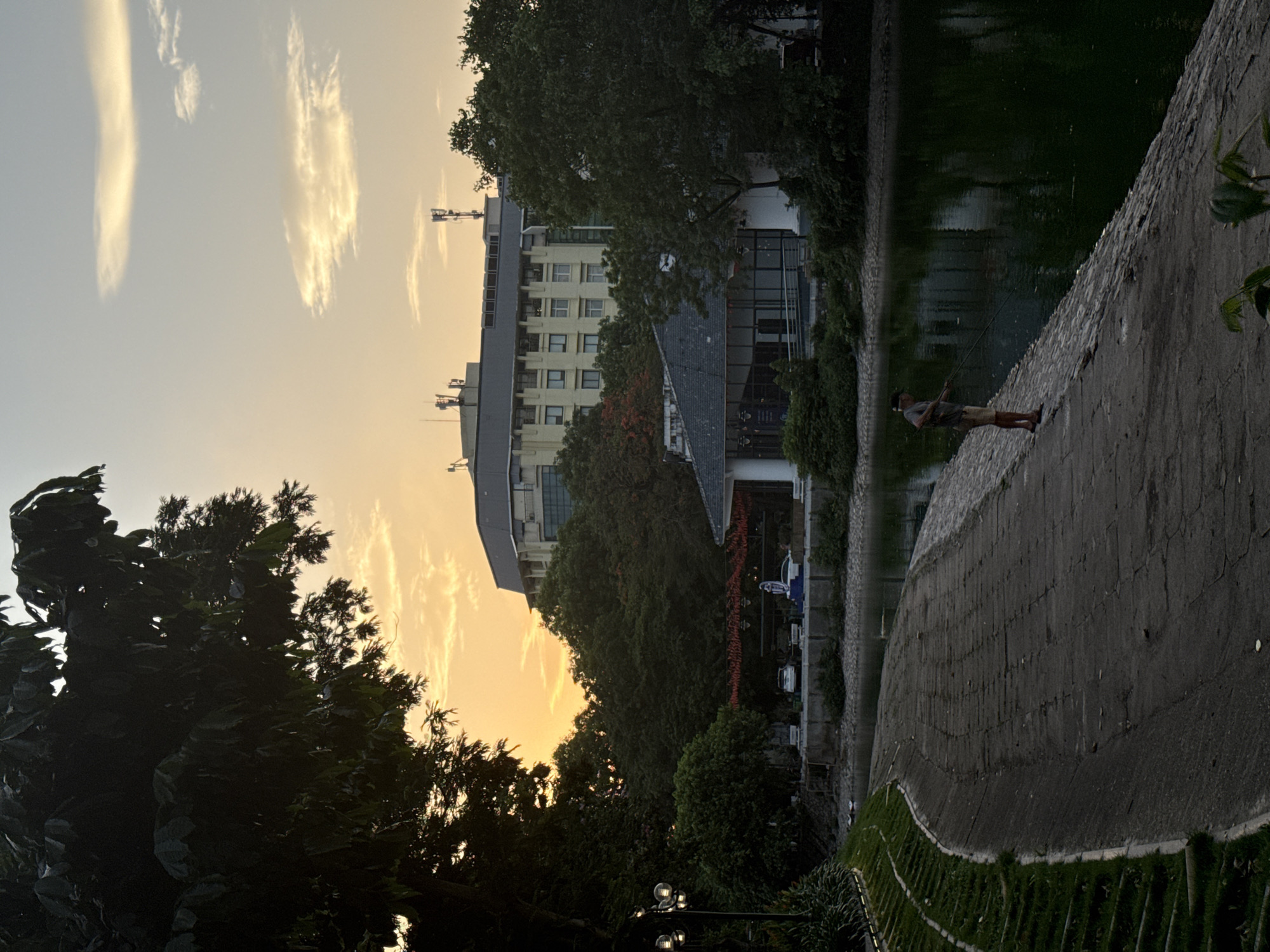 A man fishes in Hoàn Kiếm Lake in Hà Nội, Vietnam. The sun is setting. Buildings and trees are in the background.