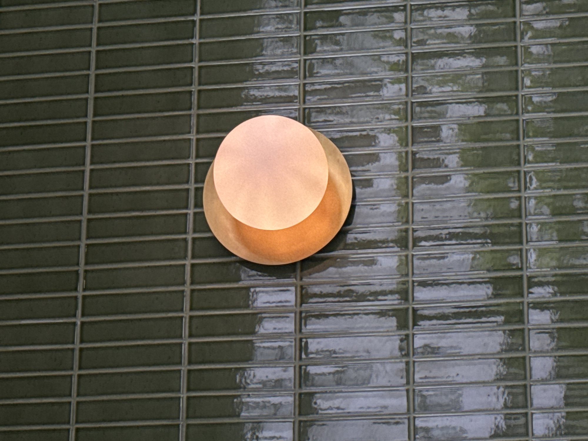 A circular, textured, orange and pinkish light fixture is mounted on a wall covered with rectangular, dark green, glossy tiles. The tiles are arranged in a vertical brick pattern with light gray grout lines. The light fixture appears to be made of two overlapping circular discs. The lower disc is a warm orange color, and the upper disc is a lighter pinkish-orange, emitting a soft glow. The tiles reflect the light and the surrounding environment, which appears to be an outdoor setting with greenery visible in the reflections. The scene is static, with no people or animals present. The image is shot from a slightly downward angle, focusing on the light fixture and the tiled wall.