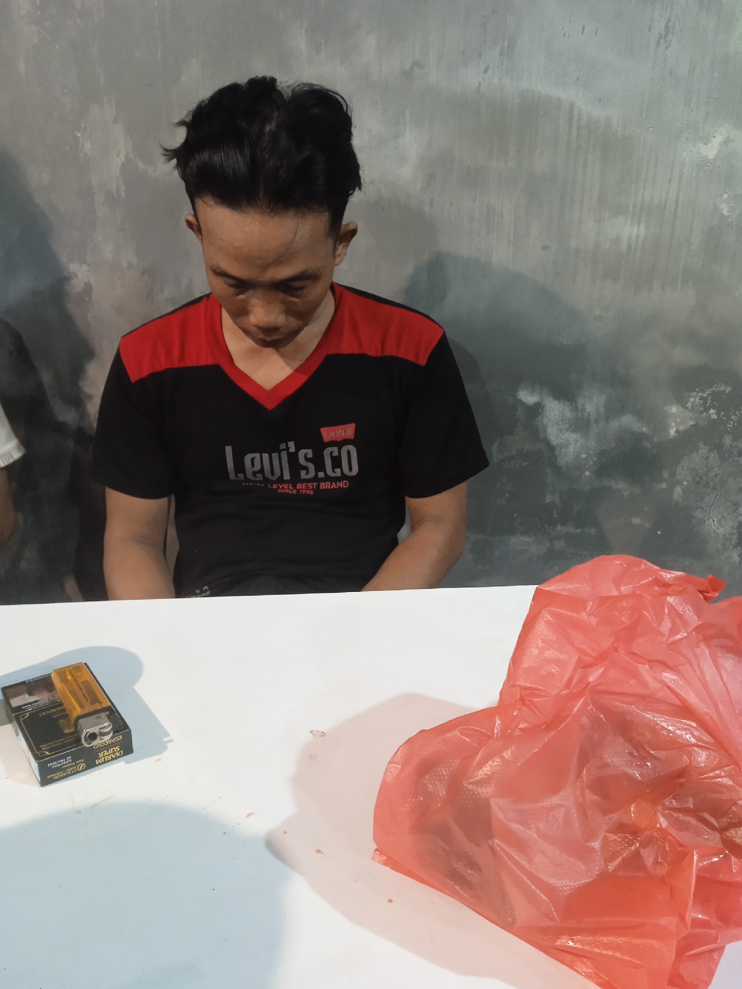 A young man with black hair and dark eyes is seated at a white table, looking downwards. He is wearing a black t-shirt with a red collar and red stripes on the shoulders. The t-shirt has the "Levi's.co" logo printed on it. Next to him, on the table, is a pack of Djarum Super Espresso cigarettes and a transparent orange lighter. To the right of the cigarettes, a crumpled red plastic bag is visible. The background consists of a rough, textured gray wall. The overall scene appears to be indoors, likely in a modest dwelling or a room in Semarang, Indonesia. The lighting suggests it is artificial light, possibly from a fluorescent bulb or overhead lamp.