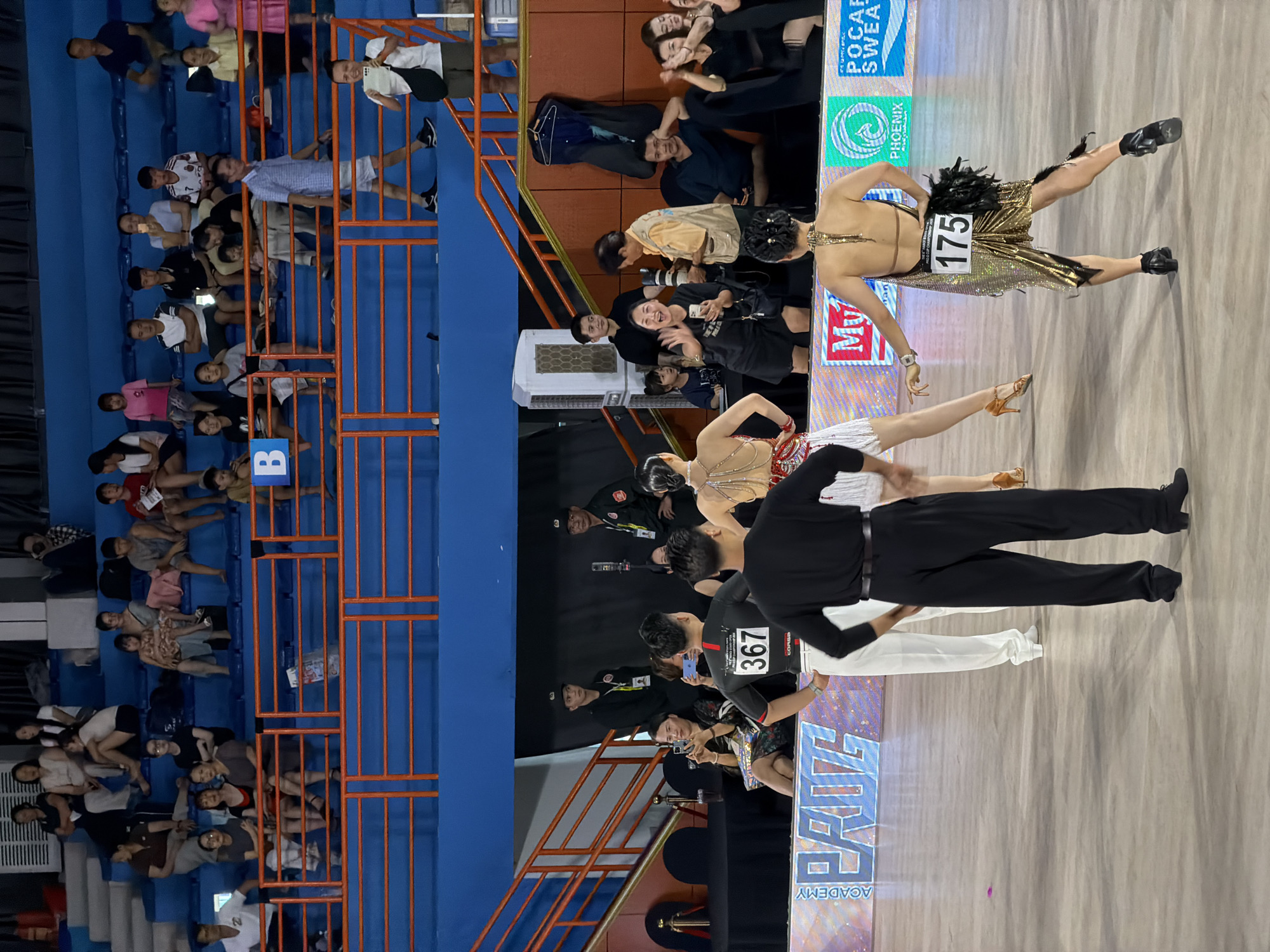 The image depicts an indoor ballroom dancing competition taking place in Hà Nội, Vietnam. The scene is a large, brightly lit hall with rows of blue stadium seating filled with spectators. In the foreground, several couples are actively dancing on a polished wooden floor. One couple, dressed in black and white formal attire, stands in the center, facing away from the camera. Another dancer, wearing a shimmering gold dress with the number 175 on the back and a black feather accent, is positioned to the right, her back to the viewer. The background shows more spectators in the stands, some of whom are taking photos with their phones. Various banners and signage are visible along the edge of the dance floor, indicating sponsors and event details. The overall atmosphere suggests a lively and competitive event.