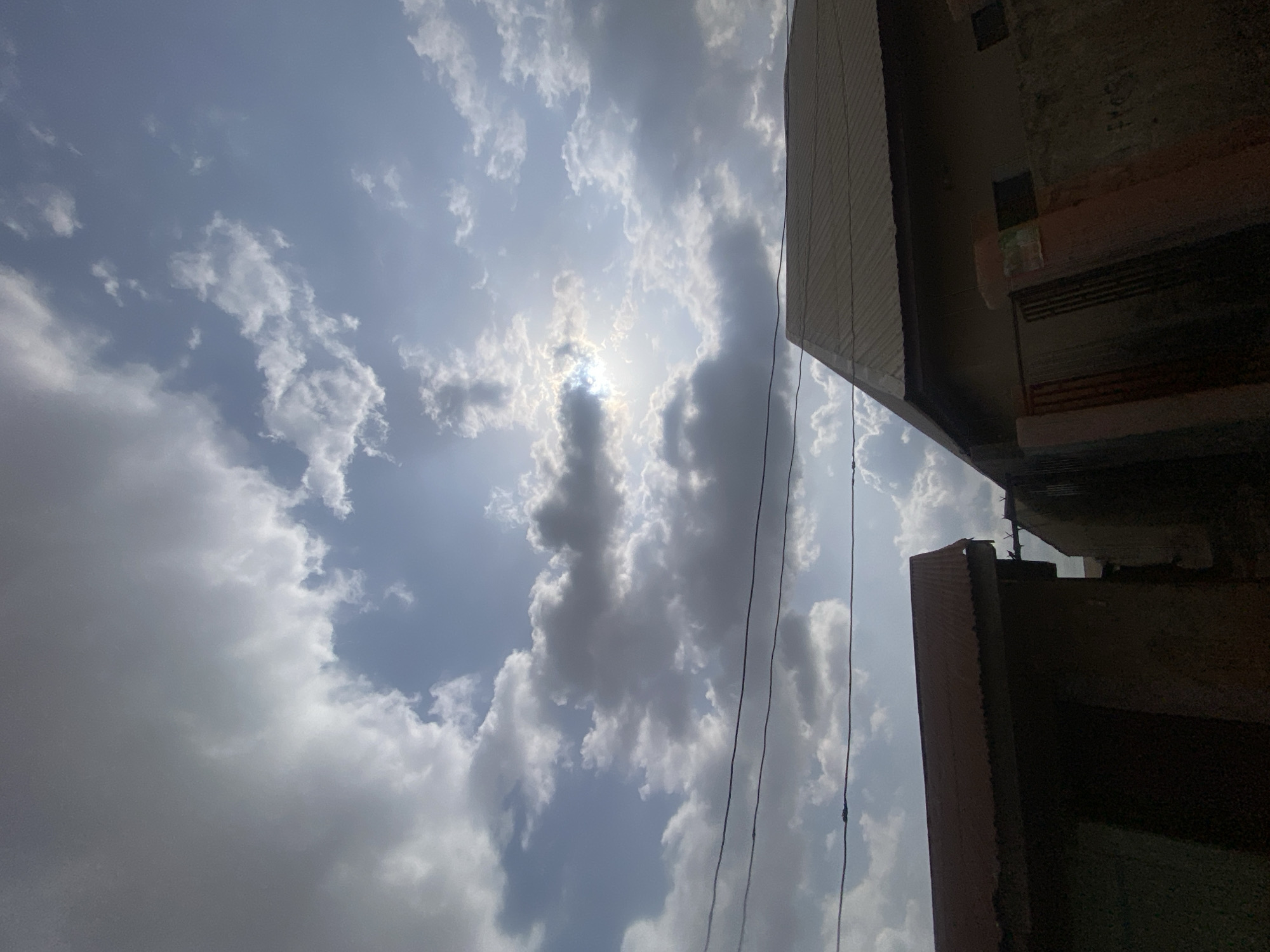 The image is a low-angle shot of the sky, predominantly filled with white and gray clouds, obscuring the sun at the center of the frame. The bright sunlight partially shines through the clouds. In the upper right and lower right corners, portions of a building are visible, with a corrugated metal roof and a dark wall. Electrical wires stretch across the sky, intersecting the cloudscape. The location is Narayi, Nigeria, as indicated in the prompt. No people or animals are visible.