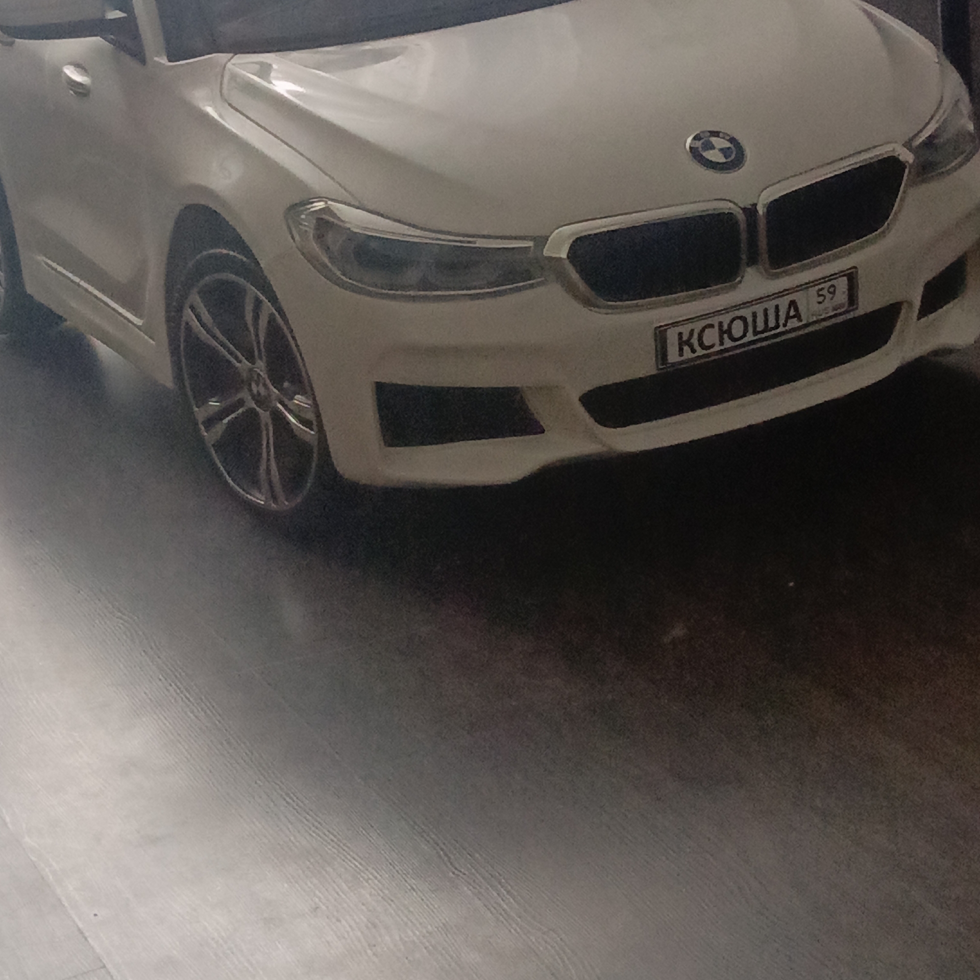 The image shows a white BMW toy car with a license plate that reads "КСЮША 59", parked on a gray wooden floor. The location is Unknown, Unknown.