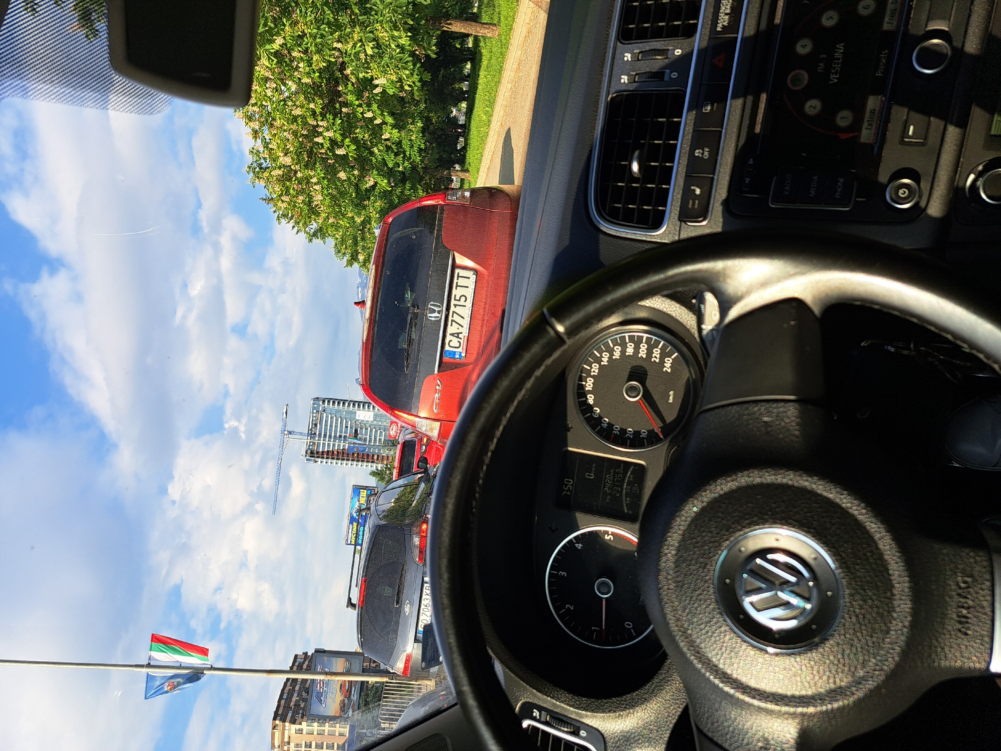 The image shows a view from inside a car, possibly a Volkswagen, with the steering wheel prominently in the foreground. Through the windshield, there is a traffic jam with several cars visible, including a red Honda CR-V with the license plate "CA 7715 TT", a grey SUV with license plate "07063 KB", and other vehicles. A Bulgarian flag and a city flag are to the left of the cars. The background includes buildings, including some modern high-rises under construction, and a blue sky with clouds. The dashboard of the car is visible. The radio is tuned to FM 3 Veselina. The odometer reading is 231753 km. The image is taken during daylight.