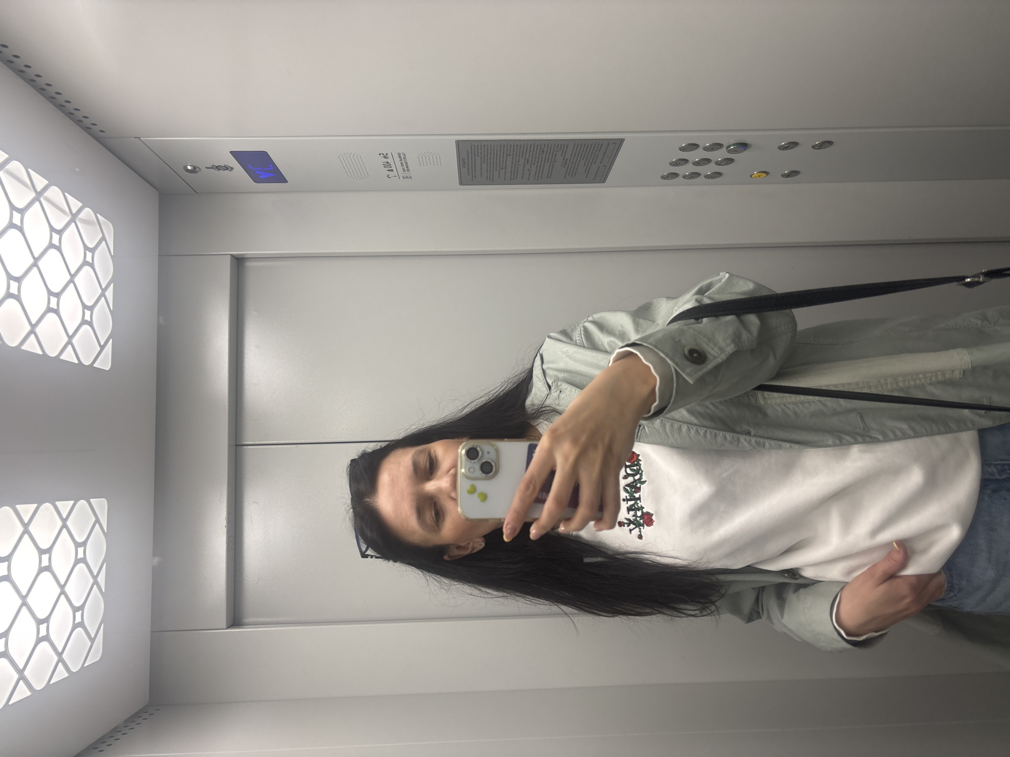 A woman is taking a selfie in what appears to be an elevator. The elevator has a gray interior, featuring patterned light fixtures on the left wall, control panels above, and a plain back wall. She is holding a smartphone to take the picture, her face partially obscured by the phone. She has long, dark hair and is wearing a light-colored jacket over a white shirt with a rose design. The location is assumed to be in the city of Chaikovsky, Russia.