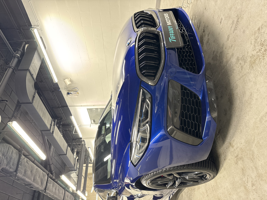 A blue BMW M8 Competition coupe is parked indoors, prominently displayed from its front-left quarter. The vehicle features a glossy metallic blue finish, black multi-spoke wheels, distinctive carbon fiber aerodynamic elements on the front bumper, and a black kidney grille. A white "TIFFANY MOTORS" license plate frame is affixed to the front bumper. The car rests on a light grey concrete floor within an industrial-style facility. The background consists of white cinder block walls and a dark grey or black ceiling with exposed rectangular ductwork, circular pipes, and numerous bright overhead fluorescent tube lights. On the right wall, a white wall-mounted unit, possibly a control panel or thermostat, with associated tubing, is visible, alongside a white speaker-like object. No individuals are present, and no specific activity is depicted. The "TIFFANY MOTORS" branding indicates the scene is located at a Tiffany Motors facility, specifically at ul. Puławska 366, 02-819 Warszawa, Poland.