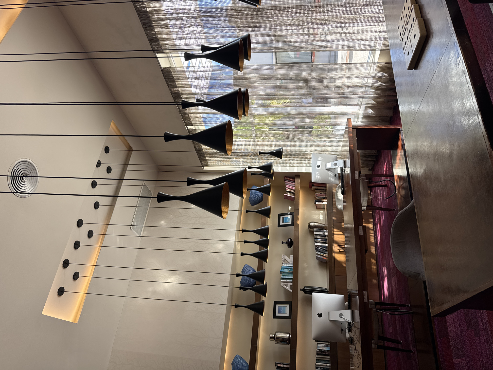 This media file captures an indoor common area, likely a modern hotel lounge, library, or co-working space. The image is taken from a slightly elevated perspective, looking across the room.
**Subjects:** There are no people visible in the scene.
**Objects:** Numerous black, cone-shaped pendant lights with golden interiors hang at varying heights from the high ceiling, forming a striking design element. A circular vent or light fixture is visible on the ceiling, along with an illuminated rectangular strip running vertically along a feature wall on the left.
In the foreground on the right, a long, dark wooden communal table is present, featuring several Apple iMac computers with their distinctive "Apple" logos visible on the back of the monitors. A wooden game board, possibly for tic-tac-toe, with square pieces, rests on the table. Partially visible grey upholstered chairs are tucked beneath the table.
Along the back wall, a tall, wooden bookshelf unit displays an array of books, decorative items such as vases, framed pictures, and unique blue woven shell-like sculptures. Some book spines show partial text, including "ALEXANDRA" and "MIA".
Large windows in the background are covered by textured, semi-sheer blinds, allowing bright natural light to filter into the room. The floor is covered with a dark red or magenta patterned carpet.
**Activity or Event:** The space appears to be unoccupied, suggesting a quiet moment before or after use. The setup is conducive to working, reading, or relaxing.
**Notable Details:** The time of day appears to be daytime, with strong sunlight casting shadows and highlights through the blinds. The overall aesthetic is contemporary and clean, with a focus on functional design and stylish decor. The combination of natural light and artistic pendant lighting creates an inviting atmosphere.
**Location Cues:** While the context provided is Playa del Carmen, Mexico, there are no specific visual cues within the image itself to confirm this location.