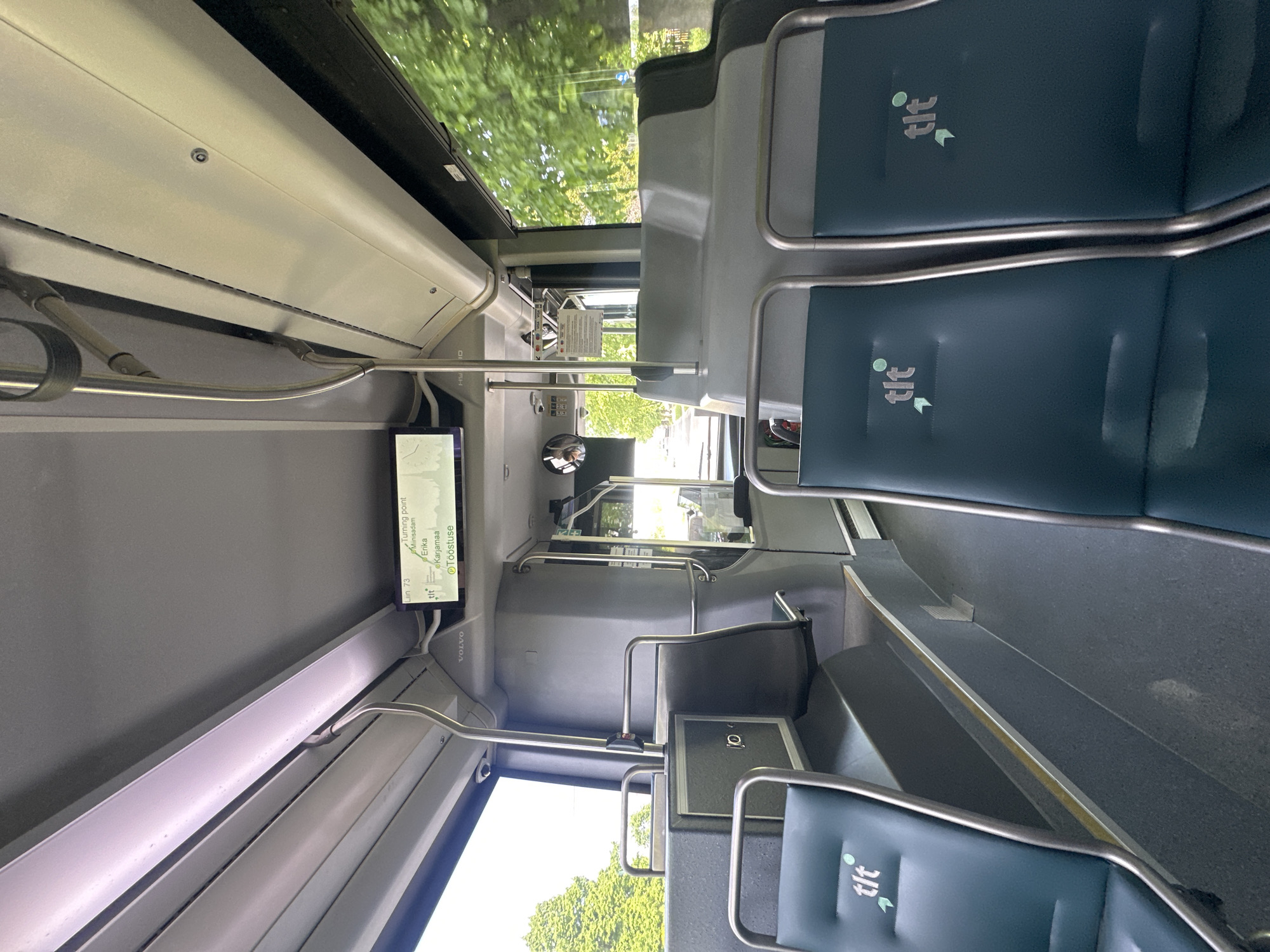 The image captures the interior of a modern public transport bus, likely in Tallinn, Estonia, given the "tlt" logo visible on the seats, which stands for Tallinna Linnatranspordi AS, the city's public transport company. The perspective is from a seated position, looking forward and slightly to the right. The seats are upholstered in a dark teal material with the "tlt" logo embossed on them.
In the foreground, two seats are partially visible. The floor is dark grey and appears to be a non-slip material. A yellow stripe runs along the edge of a raised step.
Further into the bus, the aisle is visible, leading towards the front. There are grey handrails and vertical poles for passengers to hold onto. A large, grey molded plastic structure is positioned behind the seats, likely housing internal components of the bus.
A digital display screen is mounted above the driver's area, showing route information for "Liin 73" (Line 73), with destinations like "Miinisadam", "Erika", "Karjamaa", and "Töööstuse". This confirms the location as within the bus network of Tallinn.
The windows on the right side of the bus offer a view of lush green trees and foliage, indicating an outdoor environment, likely a street or road lined with vegetation. The lighting suggests it is daytime. The overall impression is that of a clean, well-maintained public transport vehicle.