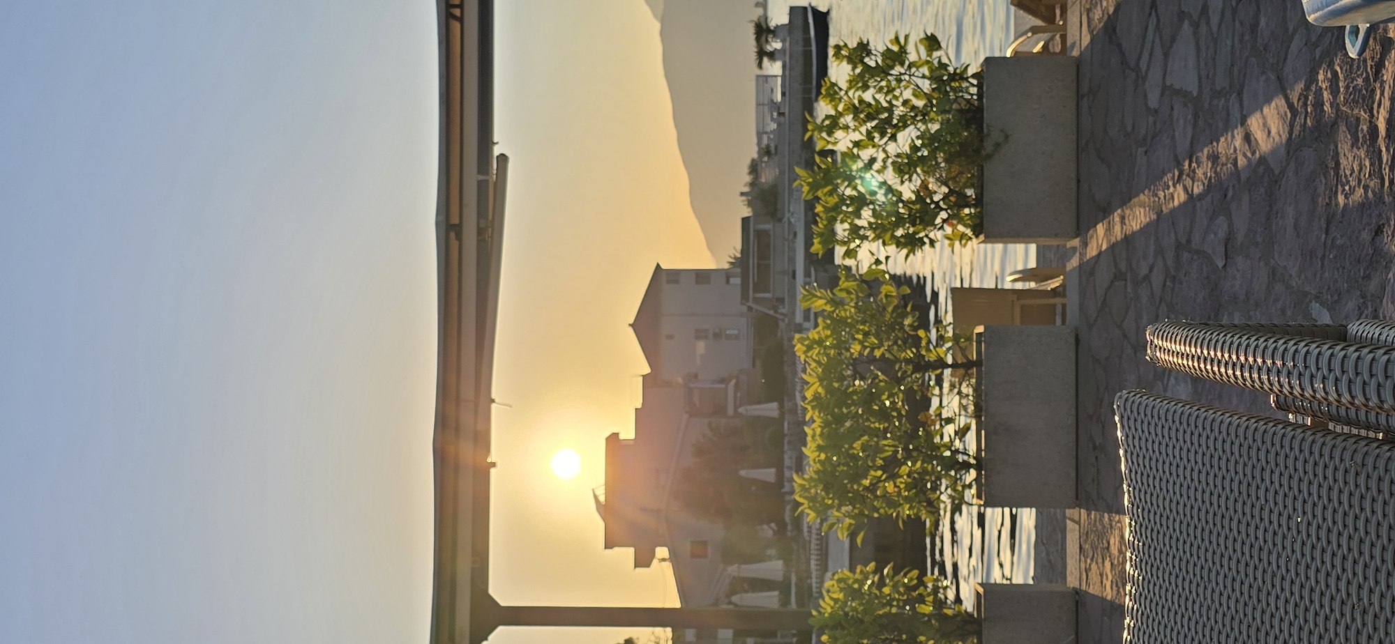 The image shows a view from what appears to be a waterfront patio in Bogišići, Montenegro. The scene is dominated by the setting sun, casting a golden glow over the landscape and the buildings. The sun is positioned behind the buildings on the shore, with a mountain range visible in the distance. In the foreground are potted trees and patio furniture, including woven chairs. The patio itself is paved with stone tiles. The water's surface reflects the golden light. A portion of a metal structure, possibly a railing or awning, is visible on the left side of the frame.