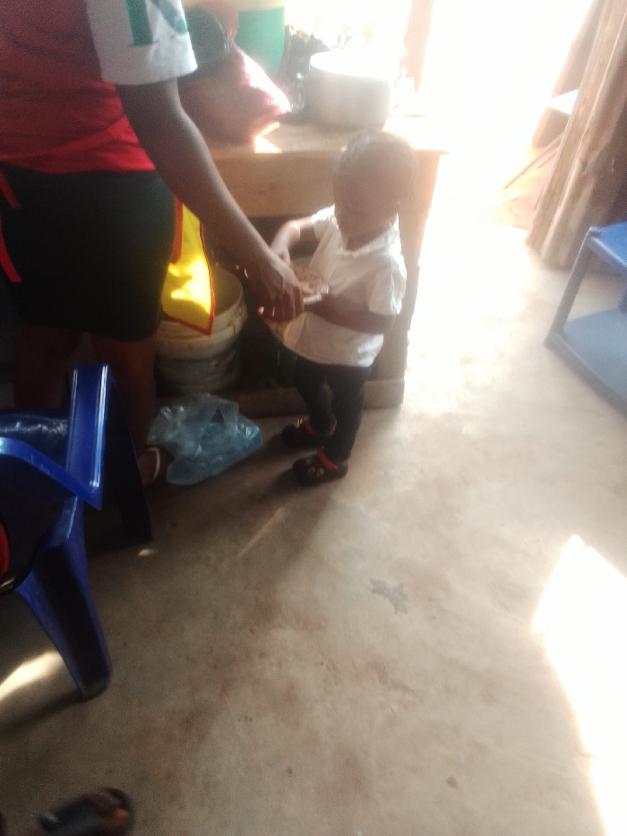 A toddler in a white collared shirt and black leggings stands next to an adult. The adult, wearing a red top with a white stripe, is assisting the child, possibly with food. A blue plastic chair is visible on the left. A plastic bag sits on the floor. Behind them, a wooden table holds items including a metal bowl. Sunlight streams in, creating strong shadows. The floor is a light tan color.