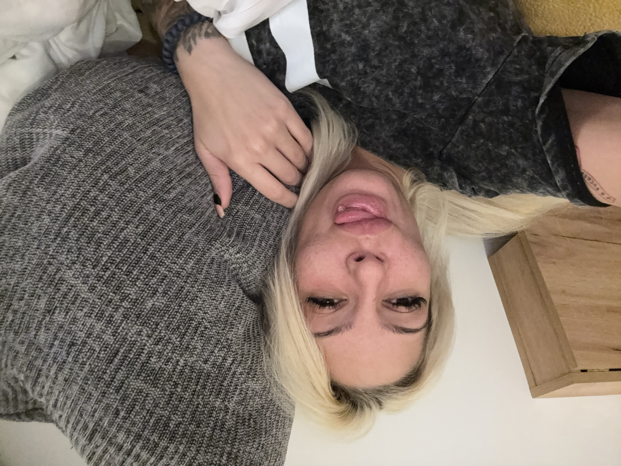 A woman with blond hair and fair skin is lying down, possibly on a couch or bed, in an indoor setting. She is sticking her tongue out towards the camera. She is wearing a gray shirt with a white stripe. Her left hand is resting on a gray knitted surface, and her left arm has tattoos. In the background, there is a light-colored wall and a wooden structure, possibly a piece of furniture. The image does not provide enough information to determine the location as Sofia, Bulgaria.