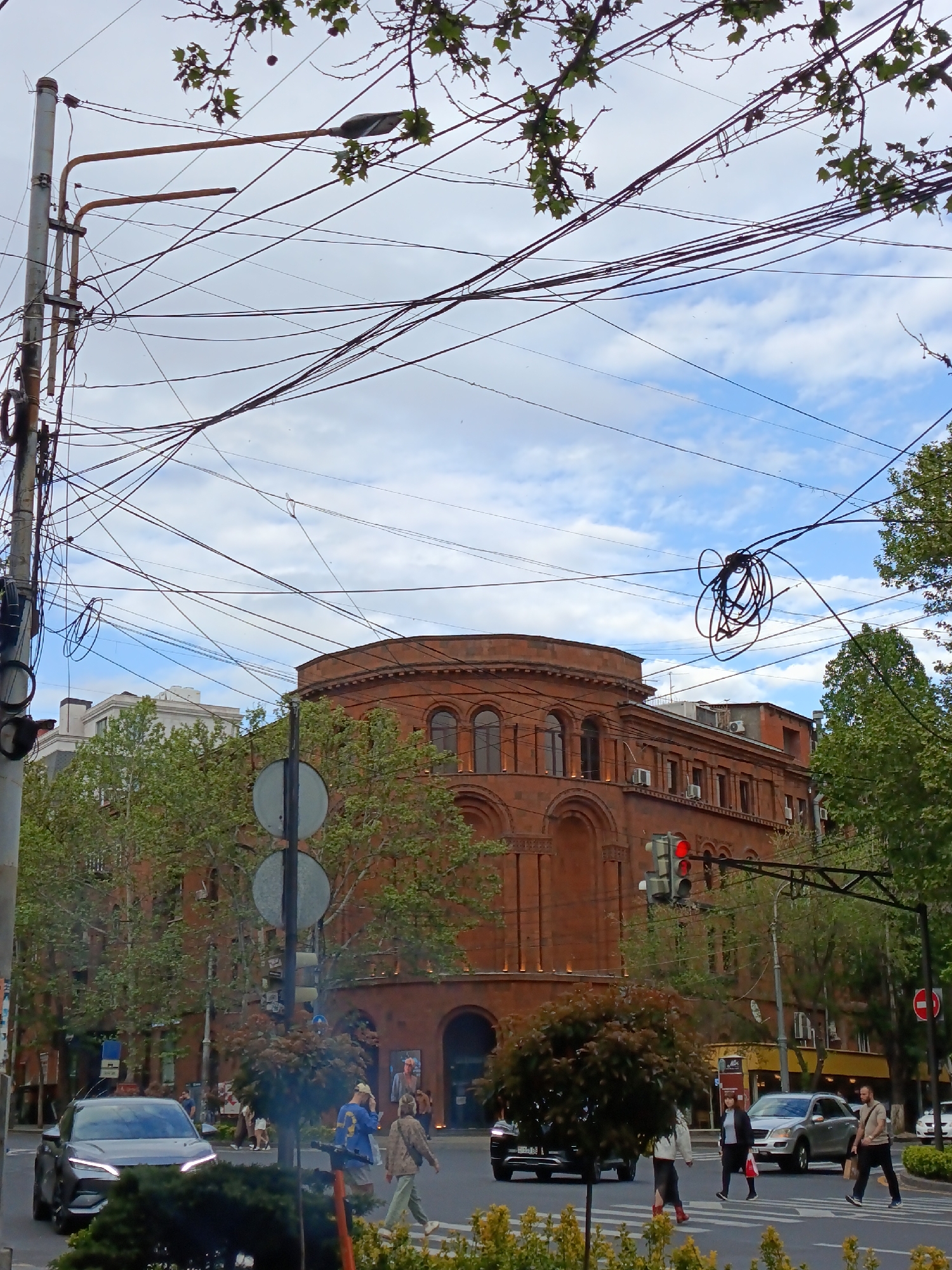 The image shows a street scene in Yerevan, Armenia, dominated by a red brick building with arched windows and a rounded top. Power lines crisscross overhead against a backdrop of blue sky with scattered clouds. In the foreground, a crosswalk is visible with several pedestrians crossing the street. Cars are stopped at the intersection, including a black sedan and a silver SUV. Traffic lights are visible, showing a red light. Trees line the sides of the street, and street signs are present. The overall impression is of a busy urban intersection in a European city.