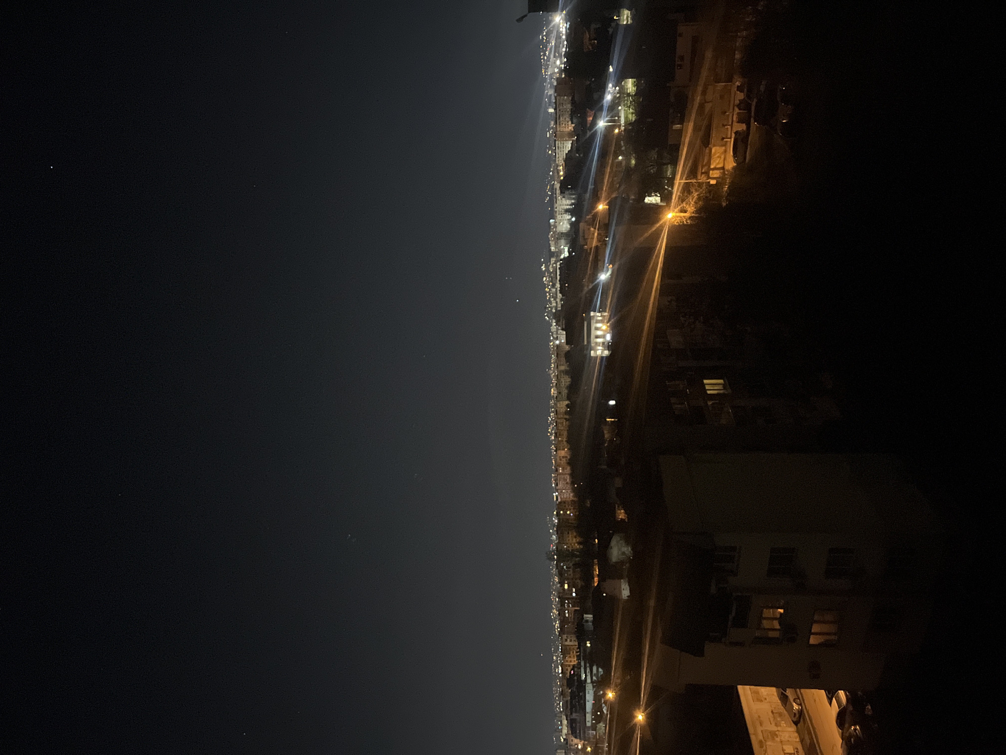 The image depicts a night cityscape. The city is likely Sofia, Bulgaria, based on the location implied in the prompt. The photo appears to have been taken from an elevated position. A dark, open area, potentially a body of water or large park, occupies a significant portion of the left side of the frame. The right side showcases the city with illuminated buildings, streetlights, and roads. The lighting suggests a typical urban setting at night. There are no visible people in the shot. The focus is on the built environment and urban landscape after dark.