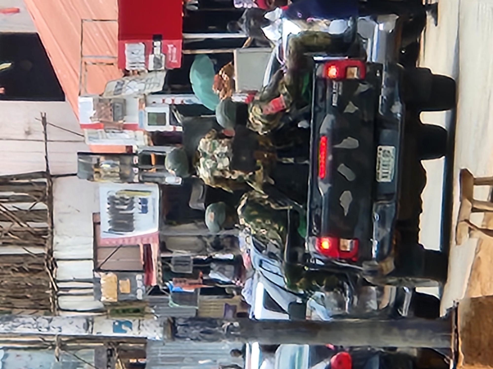 The image depicts an urban street scene, likely a commercial or market district, in the city of Unknown, Unknown. The primary subject is a black pickup truck, appearing to be a Toyota Hilux, with a dark camouflage pattern painted on its tailgate and lower rear sides. Its brake lights are illuminated. The rear license plate displays "NIML GG".
Four individuals wearing camouflage uniforms and olive-green helmets are positioned in the open bed of the pickup truck. One soldier is seated on the far left, facing forward. Another soldier stands centrally, facing forward and slightly to the right. A third soldier is seated on the far right, also facing forward, distinguished by a red armband on their right arm. A fourth soldier is partially visible behind the central standing figure.
The street is lined with various commercial structures, appearing somewhat informal, featuring metal roofing and multiple signs. A large, bright red structure or sign is visible on the right side of the frame. In the background, a green umbrella is visible amidst the street activity. A prominent vertical pole, likely a utility or light pole, stands in the left foreground. Numerous civilians are visible in the background, walking and standing near the storefronts. A civilian in a blue shirt is visible on the far right, near the truck's rear. A small wooden stool is positioned on the ground in the bottom-center foreground. The scene is illuminated by daylight.