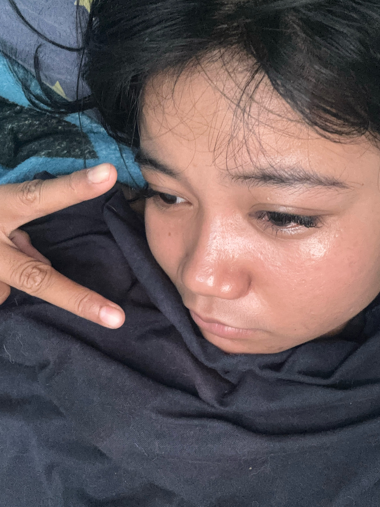 A close-up, overhead shot shows a person lying down, their head partially obscured by a black scarf or blanket. Their dark hair is spread out across a blue and black patterned blanket. The person's face is turned to the right, with their right eye visible and their left eye mostly closed, looking downwards. A hand is raised in the foreground, making a "peace" sign with two fingers extended. The person's skin appears slightly shiny, possibly from sweat or an oily complexion. The lighting suggests an indoor setting, with no specific landmarks or indications of a city location. The image is personal and informal, with no evident context beyond the immediate subject and their surroundings.