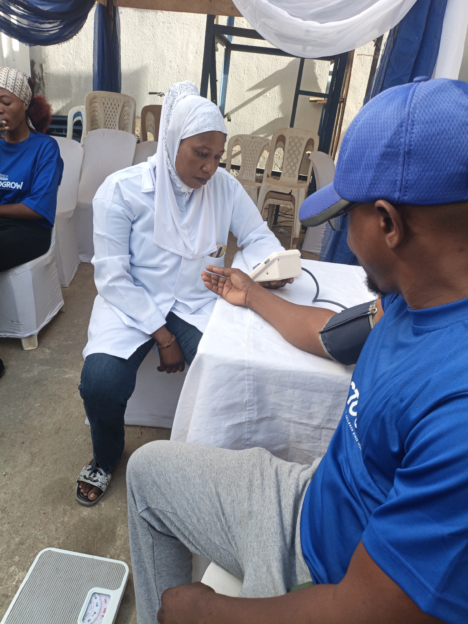 This media file depicts an outdoor community health screening event taking place during the day in Tarauni, Nigeria. The central focus is on a woman in a white lab coat and white hijab, who appears to be a medical professional, administering a blood pressure check to a man. She is seated at a white-clothed table, holding a digital blood pressure monitor, while the cuff is secured around the man's right arm. The man, seen from behind, is wearing a blue t-shirt and a blue baseball cap.
To the left of the main scene, a young woman in a blue t-shirt and a patterned headscarf is seated, looking downwards. Part of the text on her t-shirt reads "...GROW". The man's blue t-shirt also displays text, specifically "...CHILDREN SINCE 1972," suggesting an affiliation with a charitable or aid organization.
The setting is simple yet functional, featuring a temporary canopy made of white and blue fabric overhead. Several white plastic chairs, some draped in white fabric covers, are arranged around the area. A textured, light-colored wall forms the background. The ground is concrete. In the bottom left corner, a mechanical weight scale with numerical markings (0-120) is visible on the ground. The scene conveys a sense of calm and focused activity, consistent with a community health outreach effort.