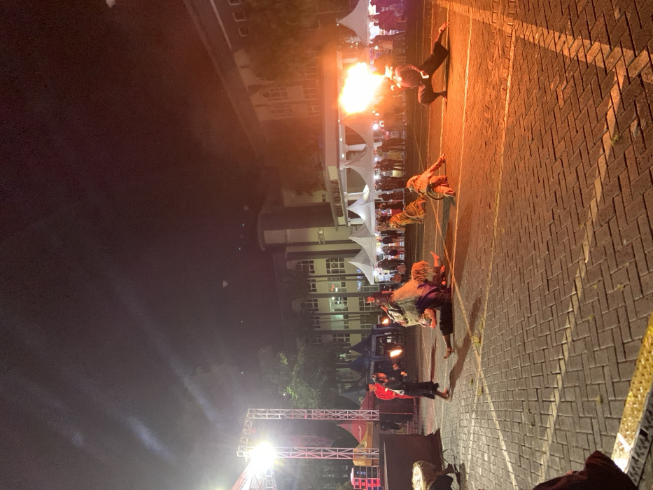 At night, a performance unfolds on a brick surface. A figure spits a burst of fire into the air, while three others crawl on the ground. Behind them, tents are set up, and people stand watching. A stage with lights and speakers is visible, suggesting a larger event. A building looms in the background.