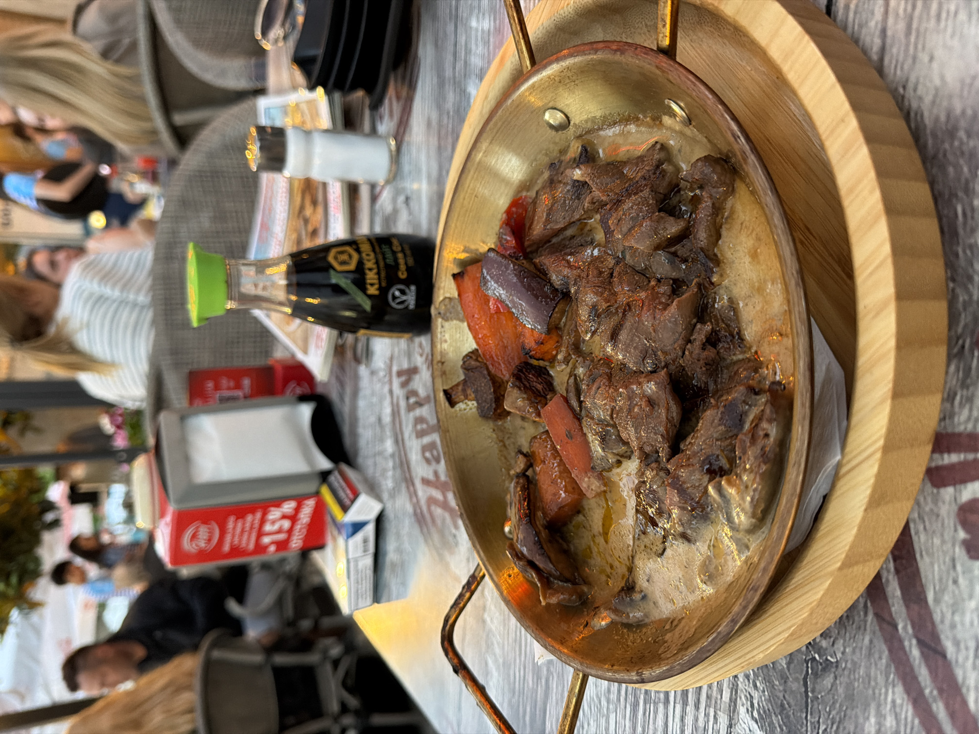 A close-up, eye-level shot captures a serving of meat and vegetables in a brass pan resting on a wooden board. The dish, possibly a stew or stir-fry, features slices of dark meat and chunks of red and purple vegetables, all coated in a light-colored sauce. The pan is set on a table with a patterned surface, part of the outdoor seating area of a restaurant. In the background, out of focus, are several people seated at tables, suggesting a bustling dining atmosphere. A bottle of Kikkoman soy sauce and a salt shaker are visible on the table in front of the dish. The scene is brightly lit, possibly by natural daylight filtered through an awning or canopy, as indicated by the warm glow on the table and the presence of what appears to be a fabric awning overhead. The overall impression is of a casual meal being enjoyed at an outdoor restaurant. This establishment is likely located in the city of Burgas, Bulgaria, given the context provided.
