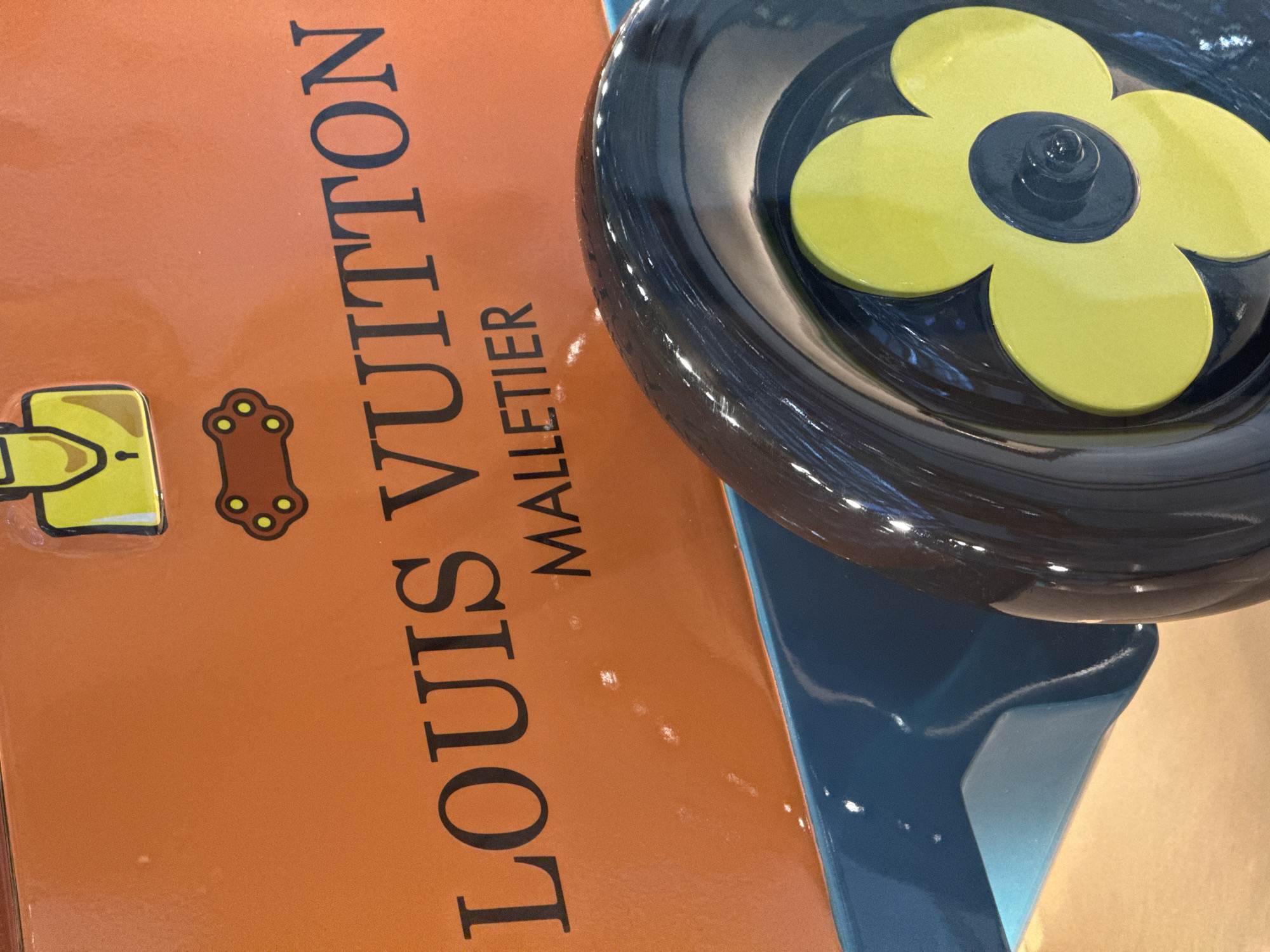The image shows a close-up of an orange object with "LOUIS VUITTON MALLETIER" printed in black. Above the text are two cartoonish brown shapes with yellow dots, one resembling a keyhole and the other a paw print. In the foreground, a black wheel with a yellow flower design is visible, attached to a teal-colored base. The scene appears to be indoors, with reflections on the surfaces suggesting artificial lighting. The image does not provide information about a specific location in Nong Prue Subdistrict, Thailand.