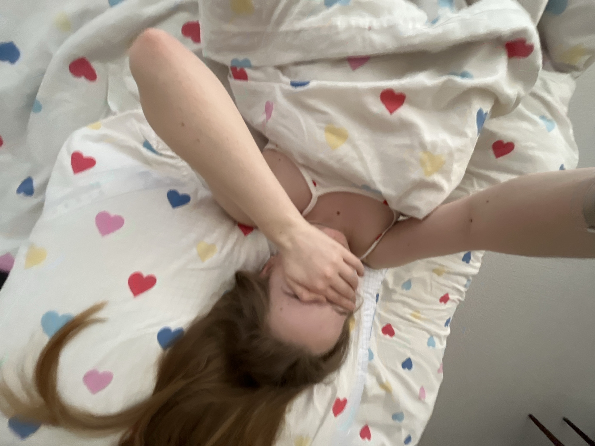 A fair-skinned person lies on a bed with white sheets and a heart pattern in blue, pink, yellow, and red. The person's face is partially obscured by their hand. Long, brown hair is visible on the left side of the frame. Part of the person's upper body is visible, clad in a white garment. The location and city are unknown.