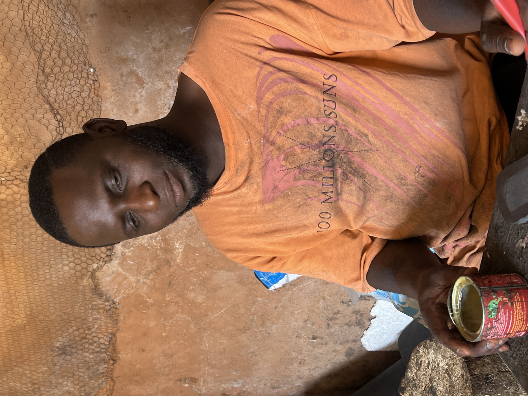 In Jos, Nigeria, a Black man with a beard and short hair is shown from the chest up. He is wearing an orange t-shirt with a graphic design and text that reads "100 MILLIONS SUNS." His head is tilted down and to the left, and he is looking downwards. In his left hand, he is holding a small, red, open tin can with a yellow substance inside. The background consists of a textured, reddish-brown wall with some netting visible at the top. To the left of the man, there is a rough wooden surface and a white, foam-like object. The overall impression is that of a working environment.