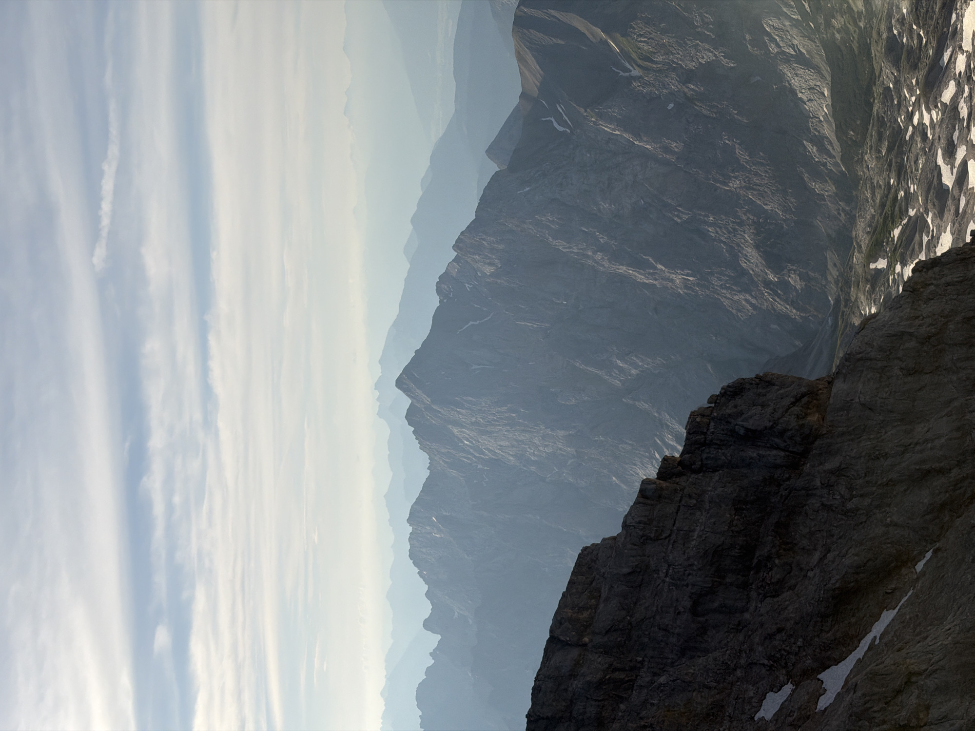 The image depicts a mountainous landscape with layers of peaks receding into the distance, indicative of a high-altitude environment. The foreground features a dark, rocky cliff face, suggesting the vantage point is from a high elevation. Below the cliff, a valley floor is visible with patches of snow amidst rocky terrain. The mid-ground and background are dominated by jagged, grey mountain ranges, softened by atmospheric haze and distance. The sky above is a pale blue with wispy, horizontal clouds. The overall impression is one of vastness, ruggedness, and natural beauty.
This scene is a view of the Zugspitze massif, the highest peak in Germany, located near Garmisch-Partenkirchen. The foreground cliff is likely a part of the Zugspitze itself, and the layered mountains in the background are other peaks within the Wetterstein range or further into the Alps. The valley floor visible could be the Höllental or a similar glacial valley. The presence of snow patches indicates a location at or above the snowline, consistent with the high altitude of the Zugspitze. No people, man-made structures, or specific actions are visible in this natural landscape.