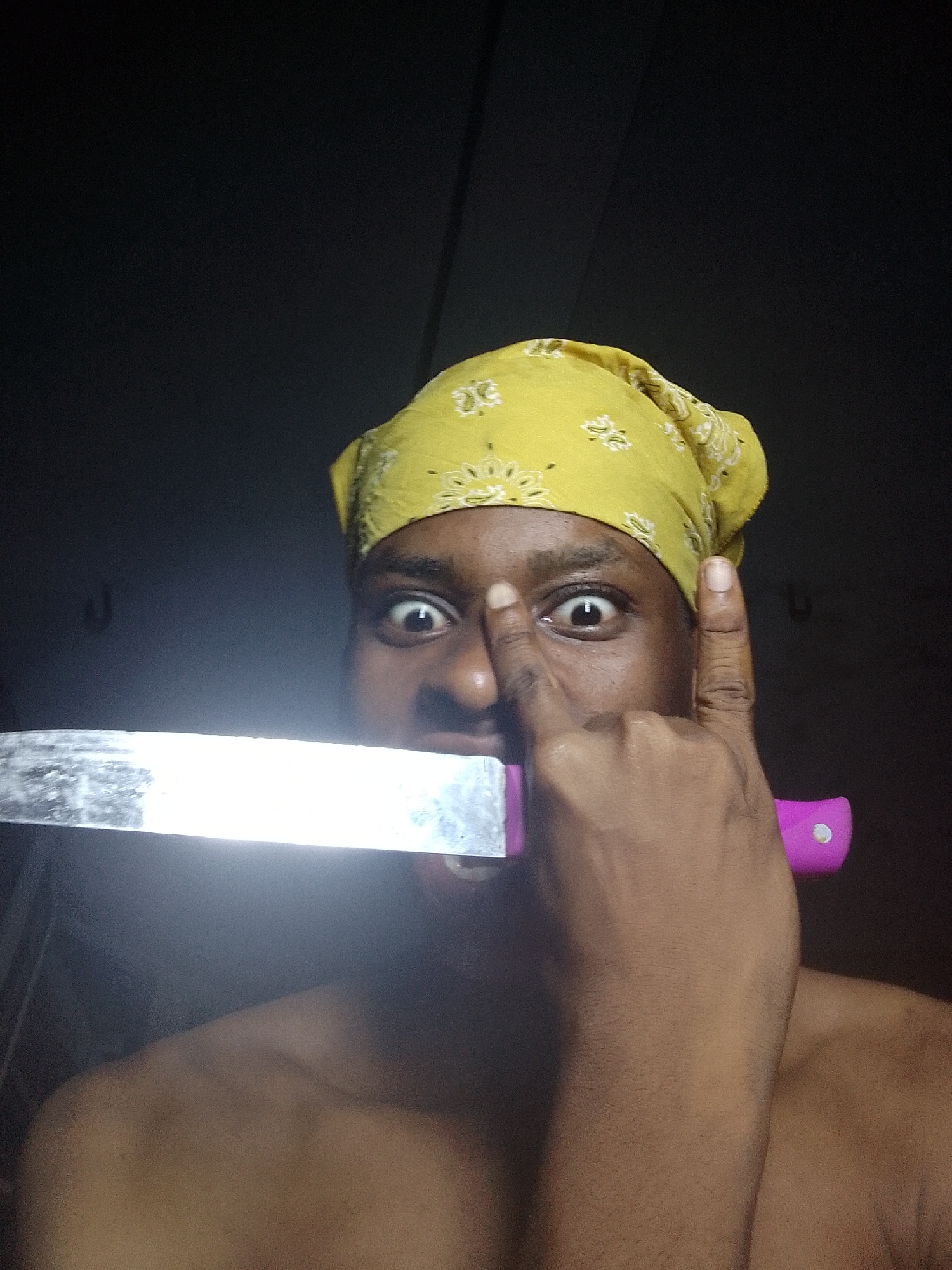 The image shows a person with dark skin wearing a yellow bandana with a floral pattern. The person is holding a knife with a shiny blade and a pink handle in their mouth. Their index finger is touching their nose, and they have a surprised expression with wide eyes. The background is dark, and there are glimpses of hooks on the wall. The location is Unknown, Unknown.