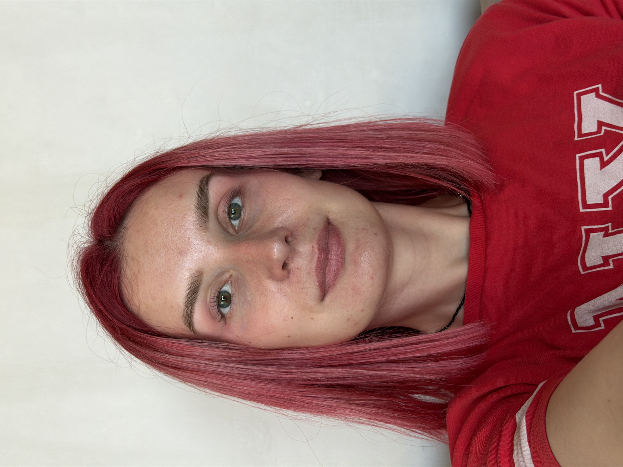 A medium shot captures a young woman with vibrant pink hair, looking directly at the camera. She is wearing a red t-shirt with white stripes on the sleeves and large white letters on the front, partially visible. Her skin is fair with visible pores and some redness around her cheeks and chin. She has light brown eyebrows and green eyes, with noticeable eye bags. Her lips are slightly parted, with a matte pink lipstick. The background is a plain, off-white wall, with no discernible features. The image appears to be taken indoors, likely in a residential setting. There is no indication of the city of Chaikovsky, Russia, being present in this image.
