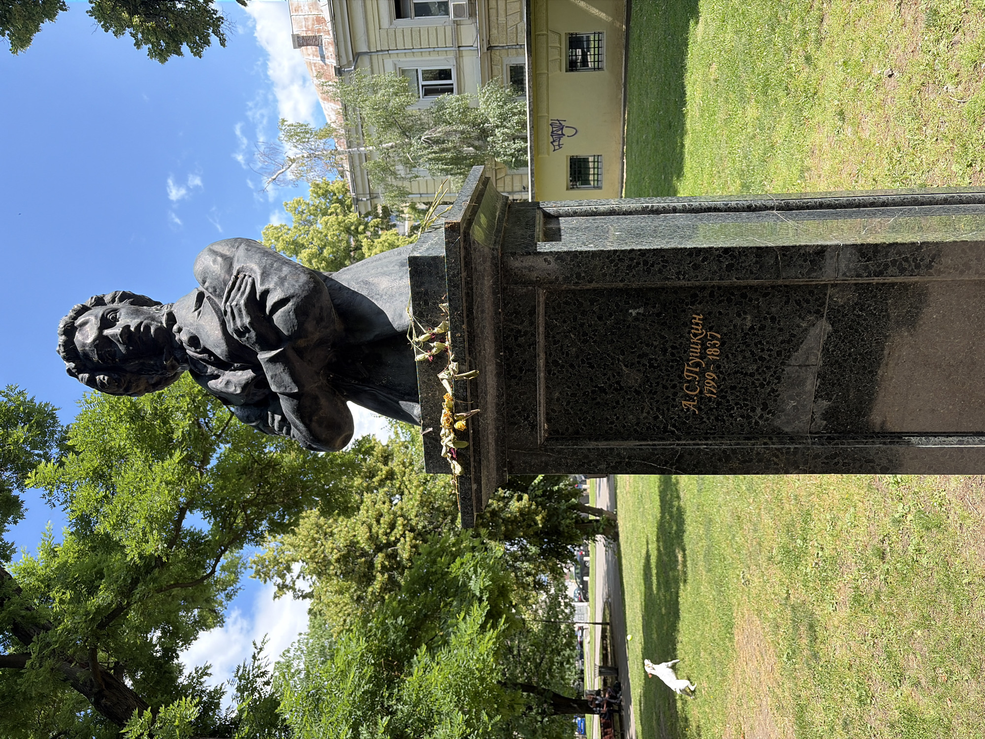 A dark bronze bust of a man with curly hair is mounted on a tall, black granite pedestal in a park. The bust depicts the man with his arms crossed and wearing a scarf around his neck. Golden Cyrillic text is inscribed on the pedestal, reading "А.С. Пушкин 1799-1837," identifying the subject as Alexander Pushkin. Several dried, wilting flowers are placed on the top edge of the pedestal.
The background of the image reveals lush green trees and a bright blue sky with scattered white clouds. Behind the trees, a light yellow building with arched windows and a red-tiled roof is visible. In the foreground, a small white dog is captured mid-air, leaping towards a yellow ball on a manicured green lawn. In the far left of the background, two individuals are seated on a park bench under the shade of a large tree, with cars parked on a street beyond them.
The photograph appears to be taken in the Borisova Gradina park in Sofia, Bulgaria, a prominent city park known for its statues and green spaces. The overall scene depicts a serene park setting with a memorial bust, a playful moment with a dog, and elements of urban architecture in the distance.