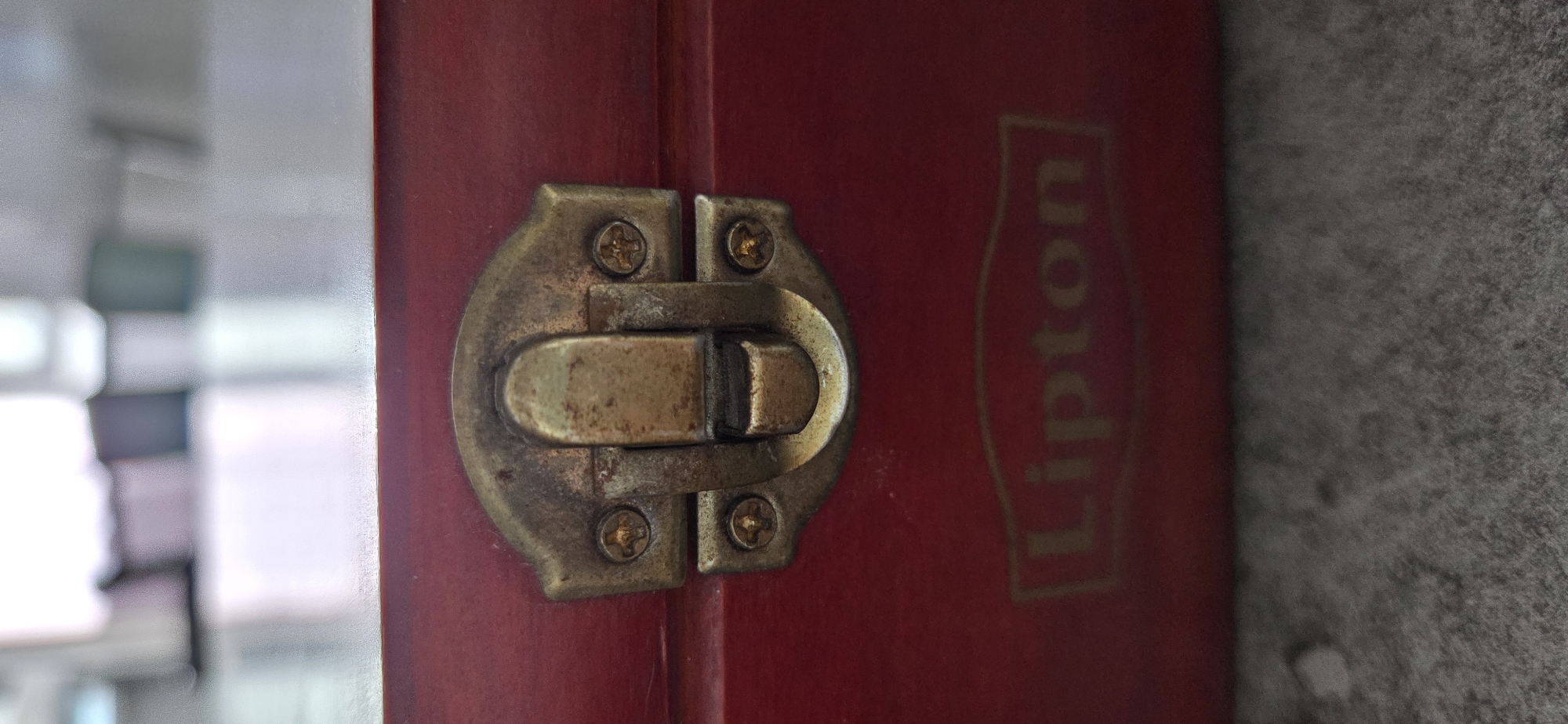 The image shows a close-up of a red wooden box with a brass latch. The Lipton logo is visible on the front of the box, embossed in gold. The box appears to be on a gray, textured surface, possibly a carpet or stone. The background is blurred and out of focus, suggesting an indoor setting. There are no people or specific landmarks visible that would indicate a location in Lipetsk, Russia.