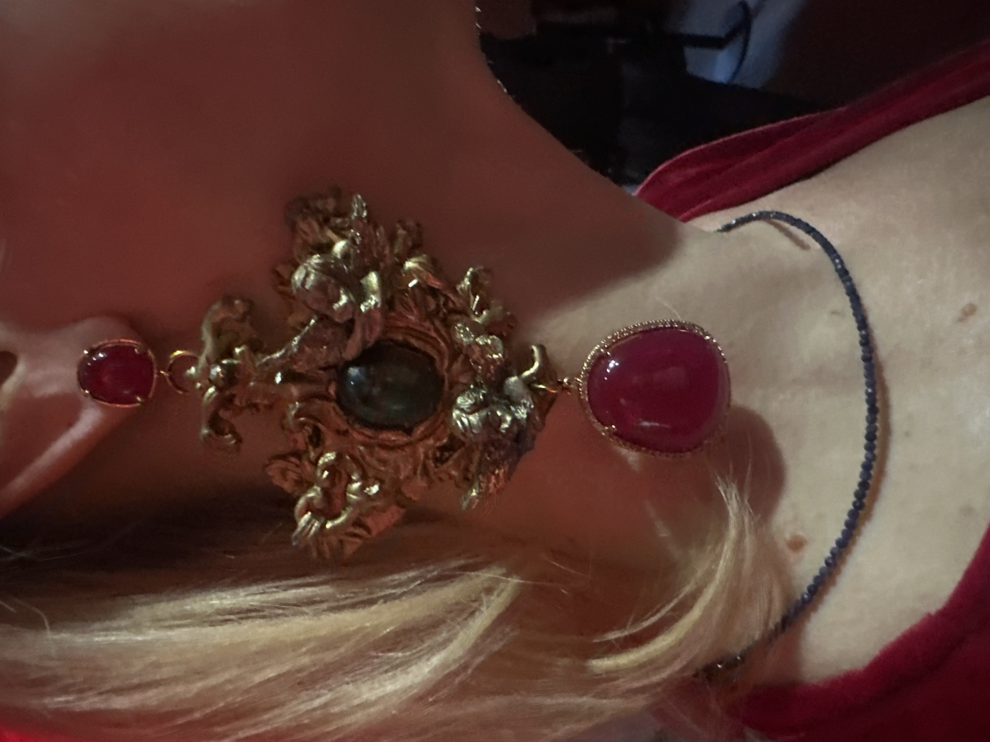 The image shows a person wearing a large, ornate gold-colored earring with multiple gemstones, including red and green stones. The earring is situated on the person's earlobe. The person also wears a delicate blue beaded necklace. They have blonde hair, and are wearing a red garment. There is no indication of the location being Riga, Latvia.