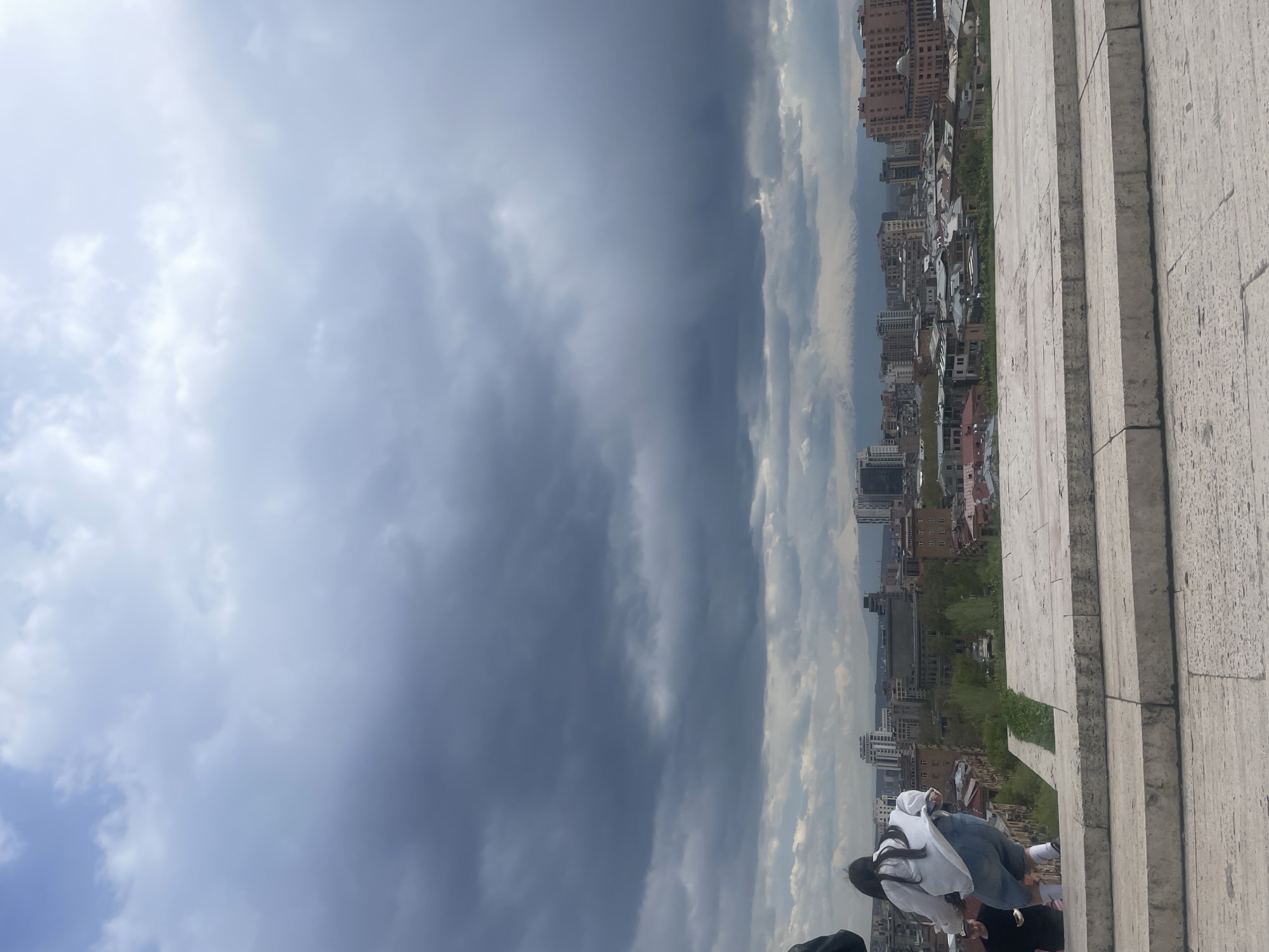 This image presents an expansive urban panorama of Yerevan, Armenia, captured from an elevated viewpoint, looking out over the city under a dramatic, overcast sky.
In the foreground, on the right side of the frame, are large, light-colored stone steps or a broad stone terrace, receding into the distance. A few individuals are visible in the bottom right corner, positioned on these steps. One person is prominently seen from behind, wearing a white long-sleeved top and blue jeans, standing and observing the cityscape. Another person, largely obscured, appears to be standing just behind them, wearing a dark top.
The midground and background are dominated by the sprawling urban landscape of Yerevan. Numerous buildings of various heights and architectural designs are visible, characteristic of a developed city. Some structures display traditional Armenian stone architecture, while others appear more modern. Patches of greenery and trees are interspersed among the buildings, adding texture to the urban environment.
The sky occupies the upper two-thirds of the image and is heavily overcast with prominent, dark grey clouds that create a moody and dramatic atmosphere. Lighter patches of sky are visible towards the upper left, suggesting diffused natural light rather than direct sunlight. The weather appears to be cloudy, possibly signaling rain or having recently rained.
The overall scene captures a moment of observation from a high vantage point, offering a broad view of the city of Yerevan under a striking sky. There is no visible text in the image. The time of day appears to be daytime, characterized by the heavy cloud cover.