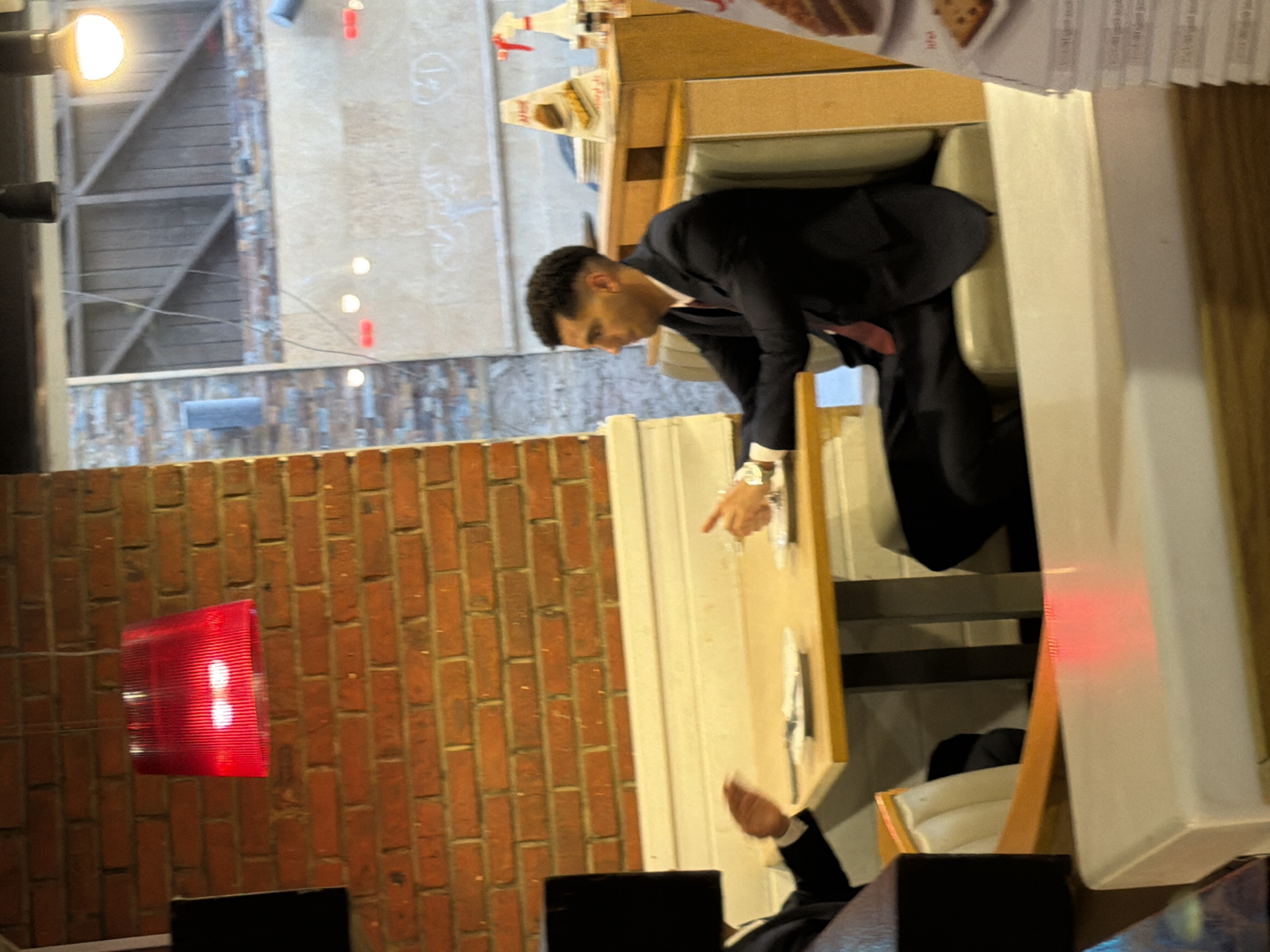 This image depicts two individuals seated at a table in what appears to be a restaurant. The man on the right is wearing a dark suit with a red tie and a watch. He is looking to his left. The man on the left is mostly obscured by a black object and is also wearing a dark suit. His right hand is visible and appears to be gesturing. The table has white plates and cutlery. The background shows a brick wall, a red lamp, and a wooden booth. A stack of menus or leaflets is visible on the right side of the frame. The overall setting suggests an indoor dining establishment. The presence of Arabic script on some of the menu items would indicate a location in an Arabic-speaking country, and given the request to specify Sharm El Sheikh, Egypt, it is likely that this is a restaurant in that city.