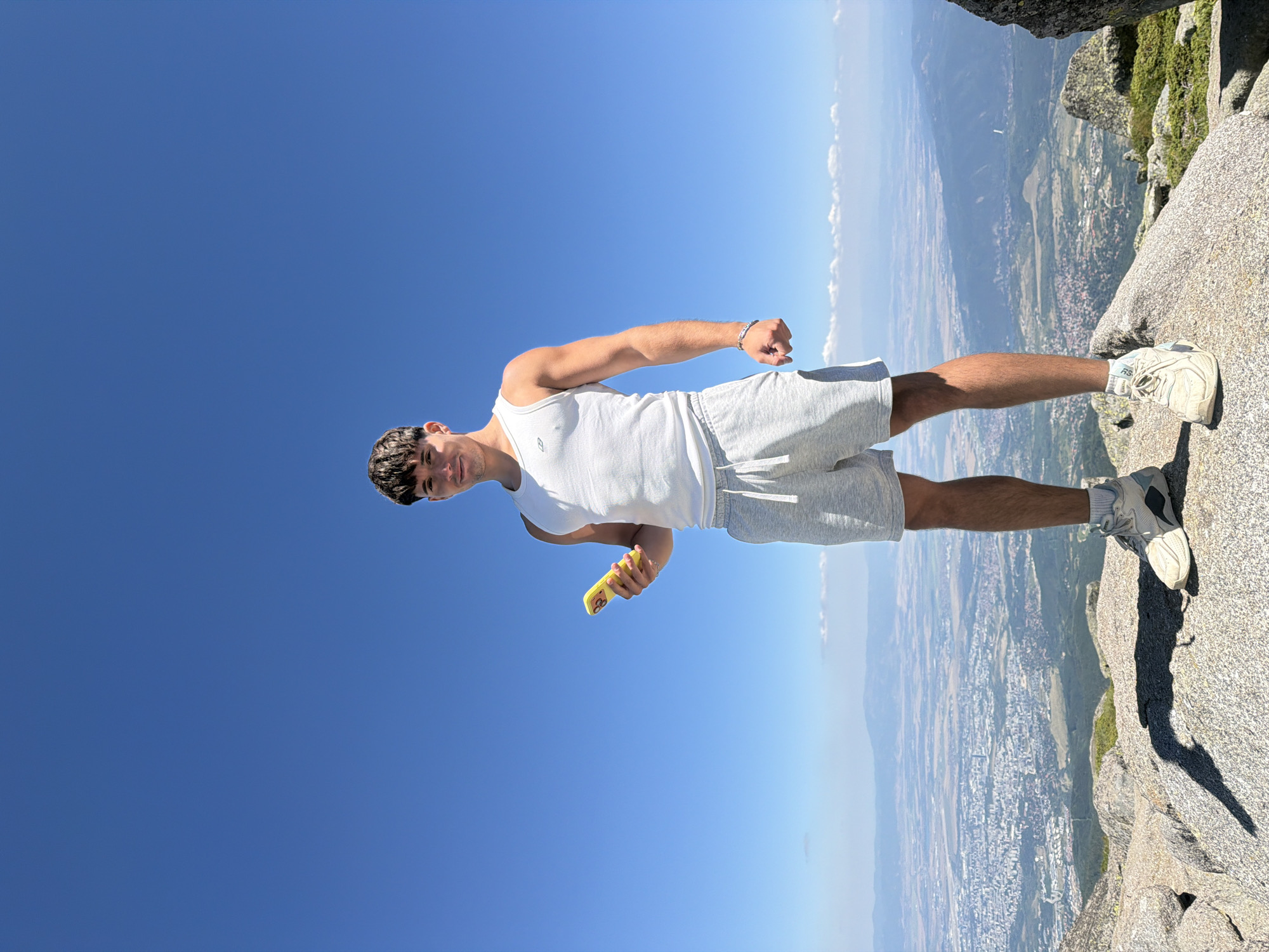 A young man stands on a rocky mountain peak, holding a yellow smartphone in his left hand. He is wearing a white tank top, light gray shorts, and athletic shoes. The background features a vast, clear blue sky and a distant city spread across a valley. The city appears to be Bistritsa, Bulgaria, with buildings and infrastructure visible from the elevated vantage point. The man is looking towards the camera with a slight smile. The lighting suggests a sunny day.
