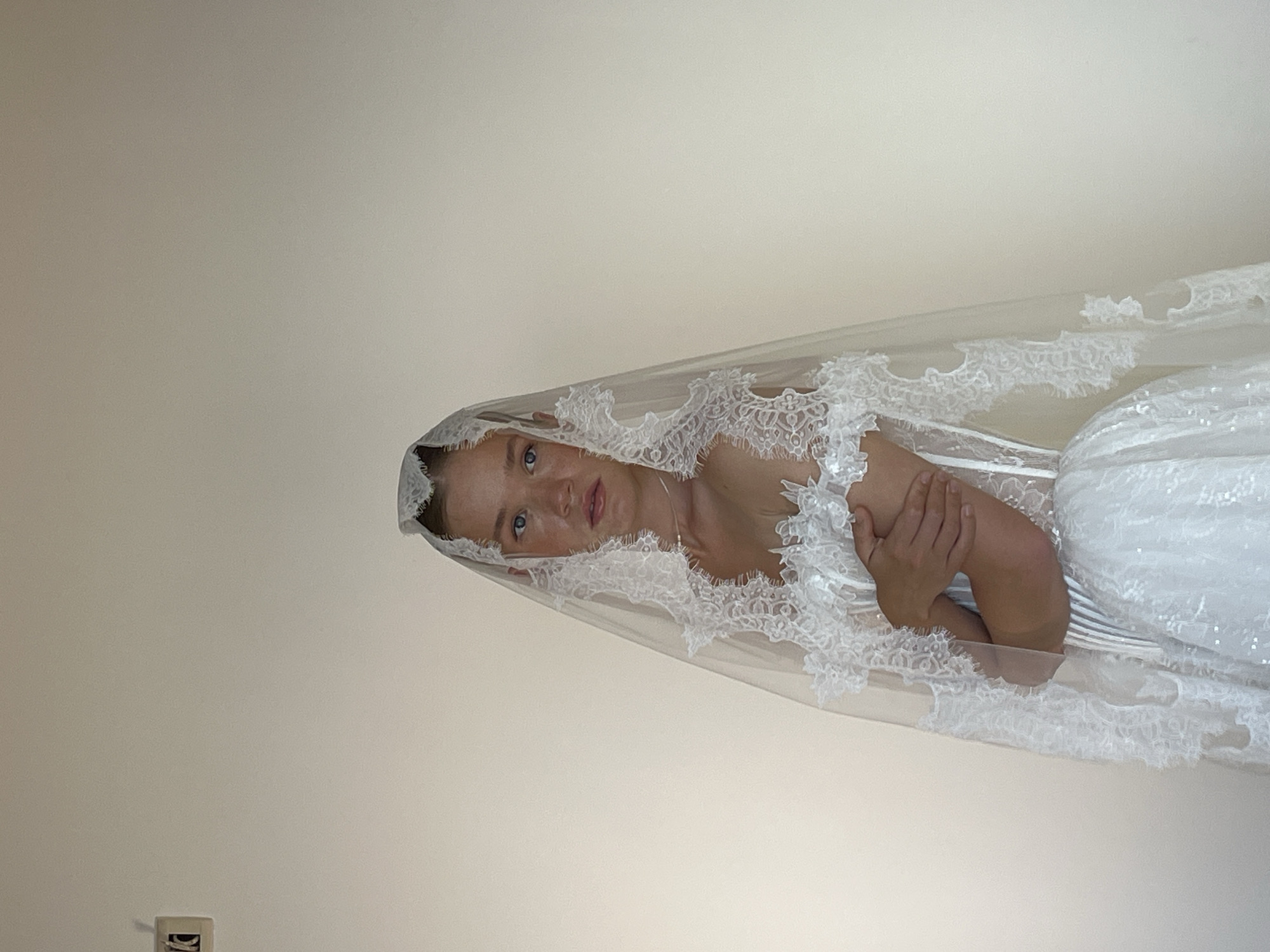 A young person with freckled skin and blue eyes, dressed in a white bridal gown and veil, stands against a plain, light-colored wall. The person is facing forward with their arms crossed. The image does not contain any discernible elements that would indicate a location in the city of Budva, Montenegro.