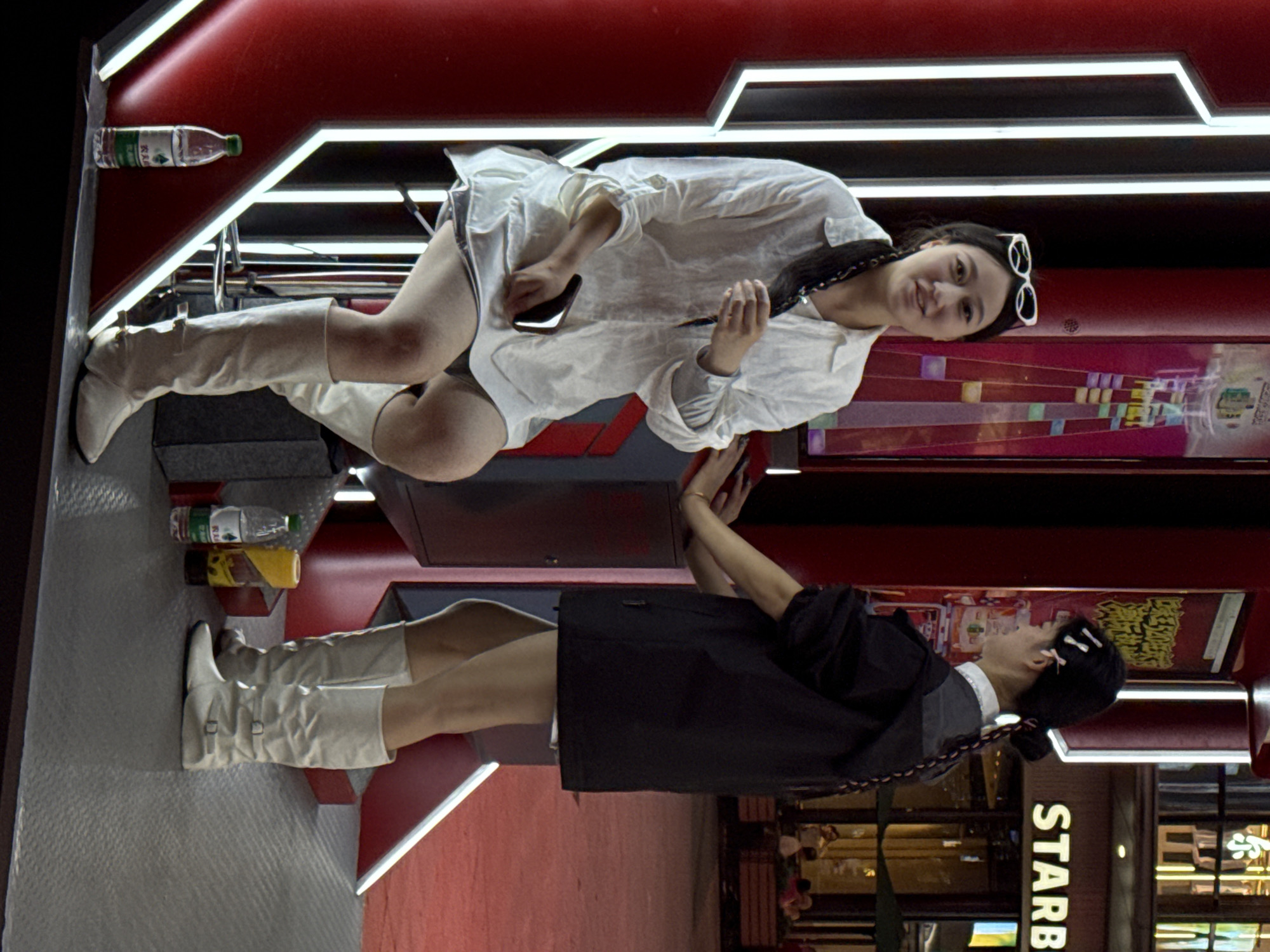Two young women are posing for a photo in front of a Starbucks in the Yuzhong District of China. The woman on the top is wearing a white dress shirt, white knee-high boots and white sunglasses, smiling and looking at the camera. The woman at the bottom is wearing a black shirt and skirt with white knee-high boots, her hair is adorned with white clips. The Starbucks logo is partially visible in the background. The scene has red lighting with geometric light fixtures above. Food and water bottles are visible on a shelf above the first woman.