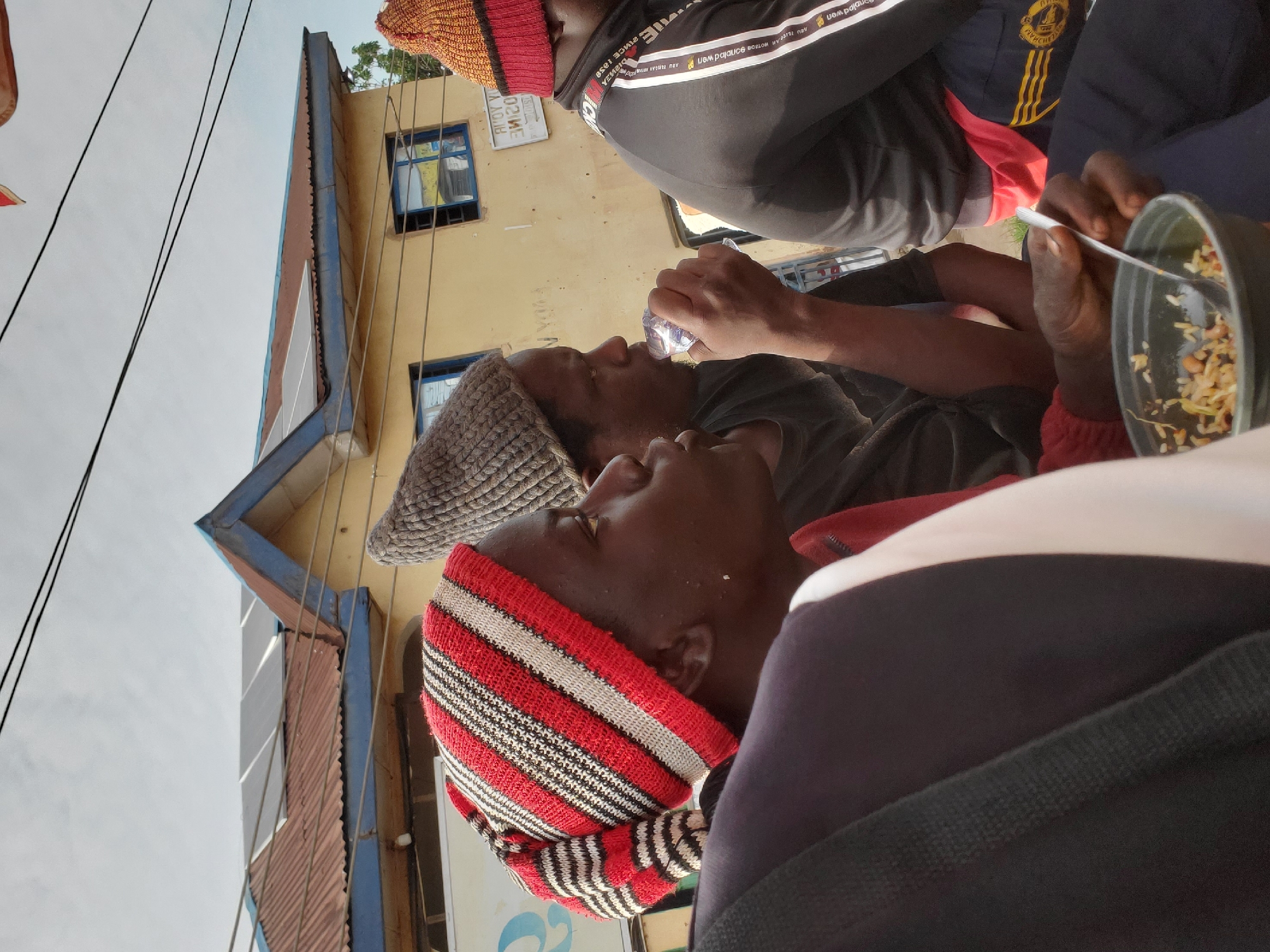 The image captures a group of individuals, likely men, in an outdoor urban setting in Gwong, Nigeria. In the foreground, a person wearing a red, white, and black striped knitted hat is seated, looking to the right. Their shoulders are visible, suggesting they are part of a larger group. To their left, another individual with a grey knitted hat is seen drinking from a clear plastic bottle. This person is looking towards the left, with their mouth near the bottle. In the background, a building with a yellow facade is prominent. It has a brown corrugated metal roof with several solar panels installed on it. The building also has blue-framed windows. Power lines are visible against a hazy sky. Further to the right, another person wearing a red and orange knitted hat is partially visible, facing away from the camera. They are wearing a black jacket with white stripes on the sleeves and possibly dark pants with yellow stripes. In the lower right corner of the image, a bowl containing rice and beans is visible, with a spoon resting in it, suggesting a meal is being consumed or prepared. The overall impression is of a candid moment, possibly documenting everyday life or a communal gathering in a Nigerian city.