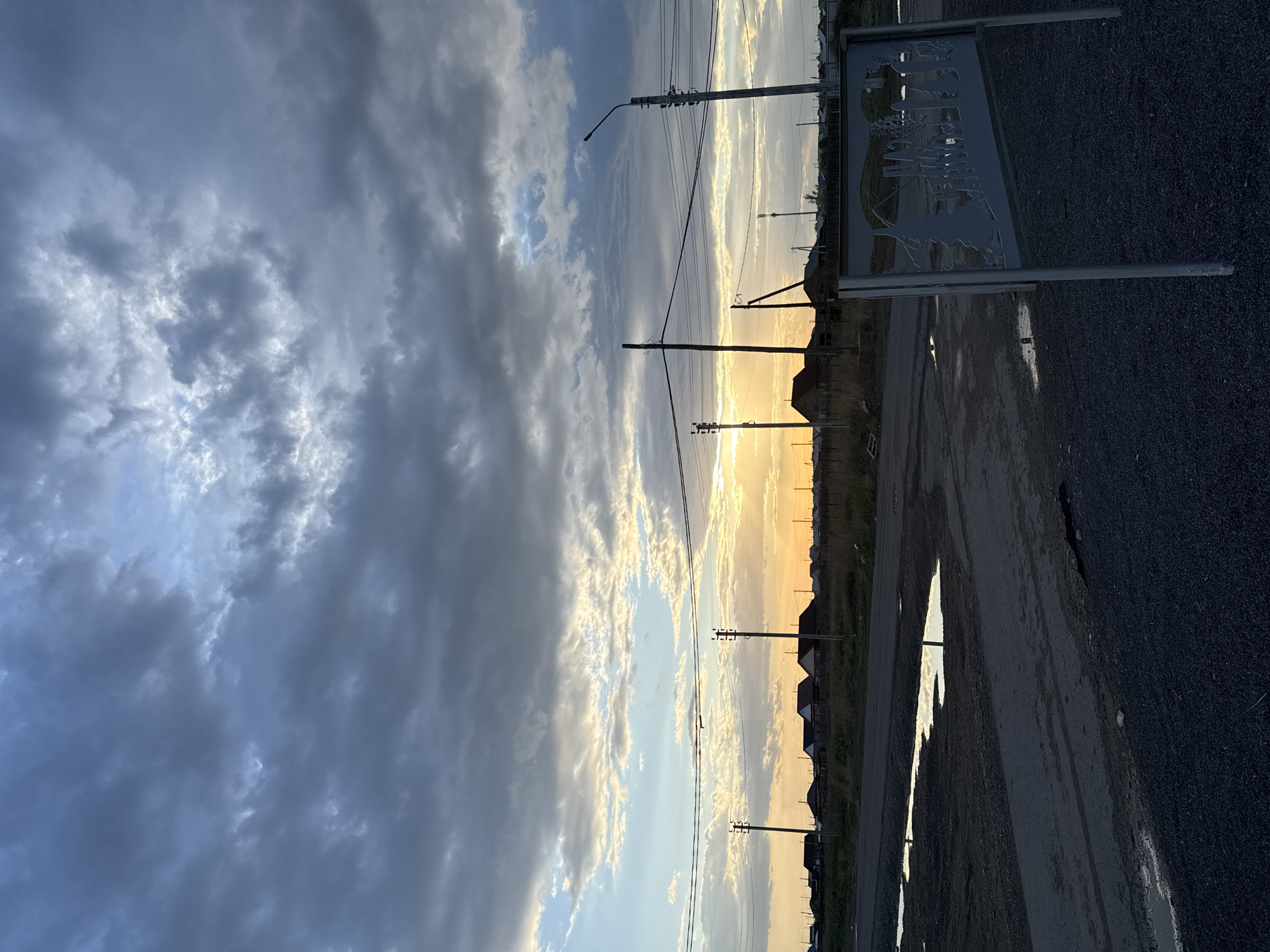 The image shows a view in Bely Yar, Russia, at dusk or dawn, characterized by an expansive sky filled with clouds. To the left, dark, dense clouds dominate, contrasting with the right side of the sky where the sun is either rising or setting, creating a bright, golden glow that illuminates the clouds.
Below the sky, a row of houses is visible along the horizon line. Power lines and utility poles run along the road, adding vertical elements to the scene.
In the foreground, a paved road stretches into the distance. Puddles on the road reflect the sunlight. On the right side of the road is a sign with metal cutout silhouettes depicting what appears to be scenes from Bely Yar and the name of the city. The ground is gravel.