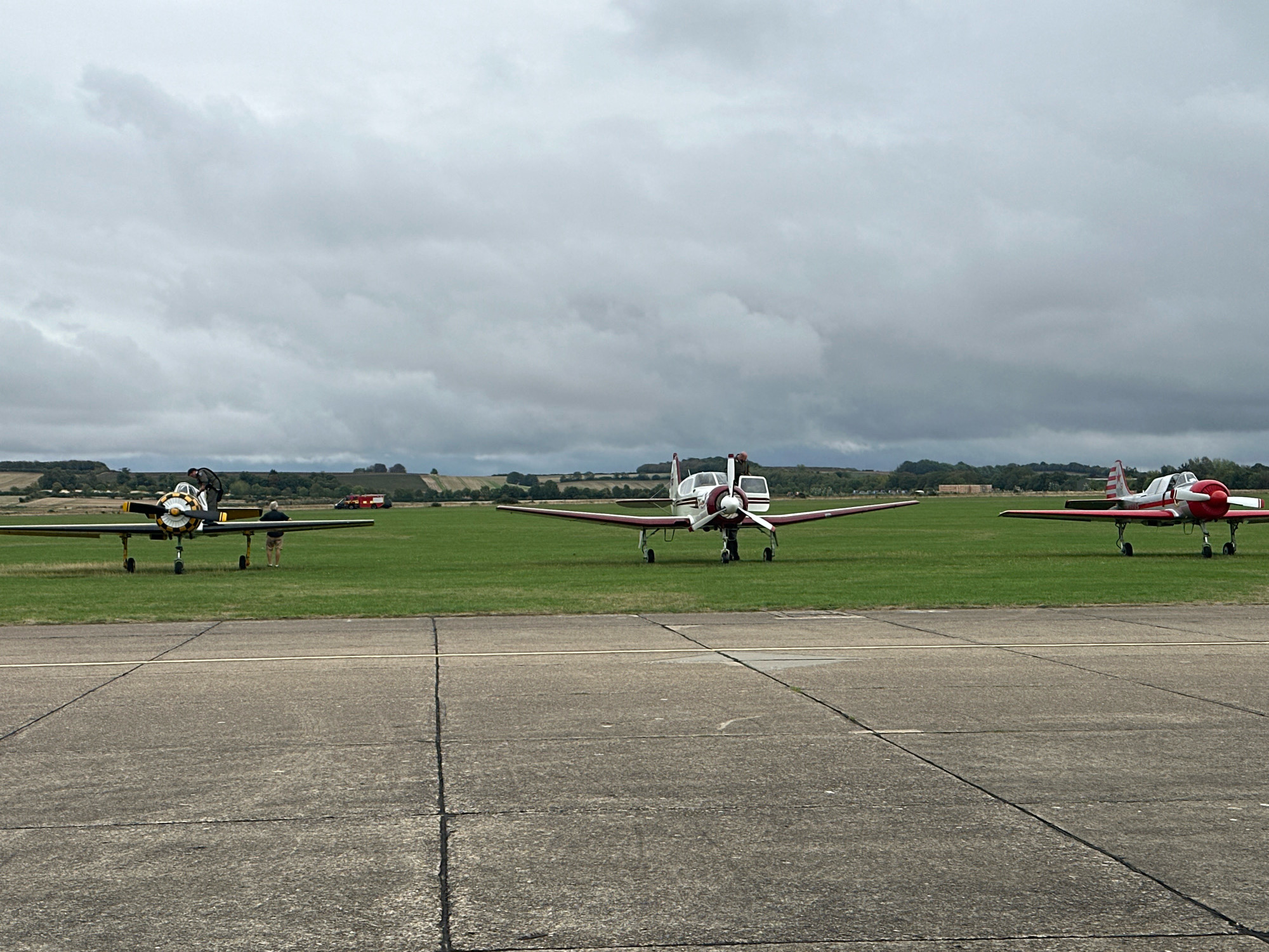 Three vintage propeller aircraft are parked on a grass airfield. The aircraft are lined up from left to right. The first aircraft on the left is yellow and black with a prominent propeller. Two people are standing near it. The middle aircraft is white and maroon with a similar propeller. The third aircraft on the right is white and red with a striped tail. A person is visible standing near it. In the background, there is a treeline and rolling hills under a cloudy sky. The foreground consists of a concrete tarmac with visible expansion joints. The scene is consistent with an airfield near Cambridge, United Kingdom.