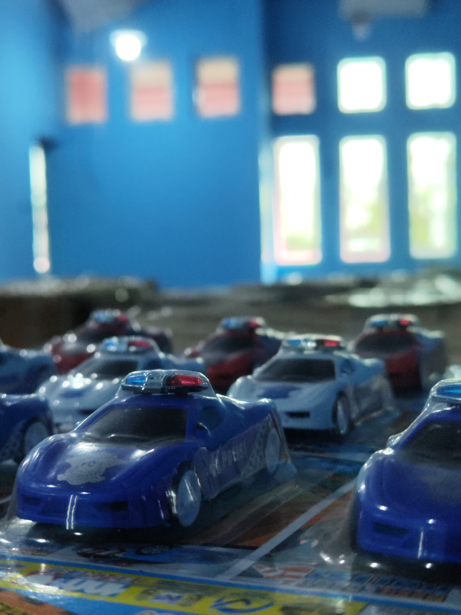 A close-up, shallow depth of field photograph captures several toy police cars lined up in what appears to be a retail display. The foreground is dominated by a blue toy police car with a silver decal and red and blue flashing lights on its roof. Behind it, several other toy police cars are visible, including a white one with red and blue lights, and others in red and blue. The cars are packaged in clear plastic, indicating they are for sale. The background is blurred, showing a blue wall with what look like windows and some artwork or signage. The overall impression is of a collection of toys presented for purchase, possibly in a toy store or market stall. The image is undated and does not provide explicit geographical context beyond the inferred location of Sragen, Indonesia, which is not visually evident in the frame.