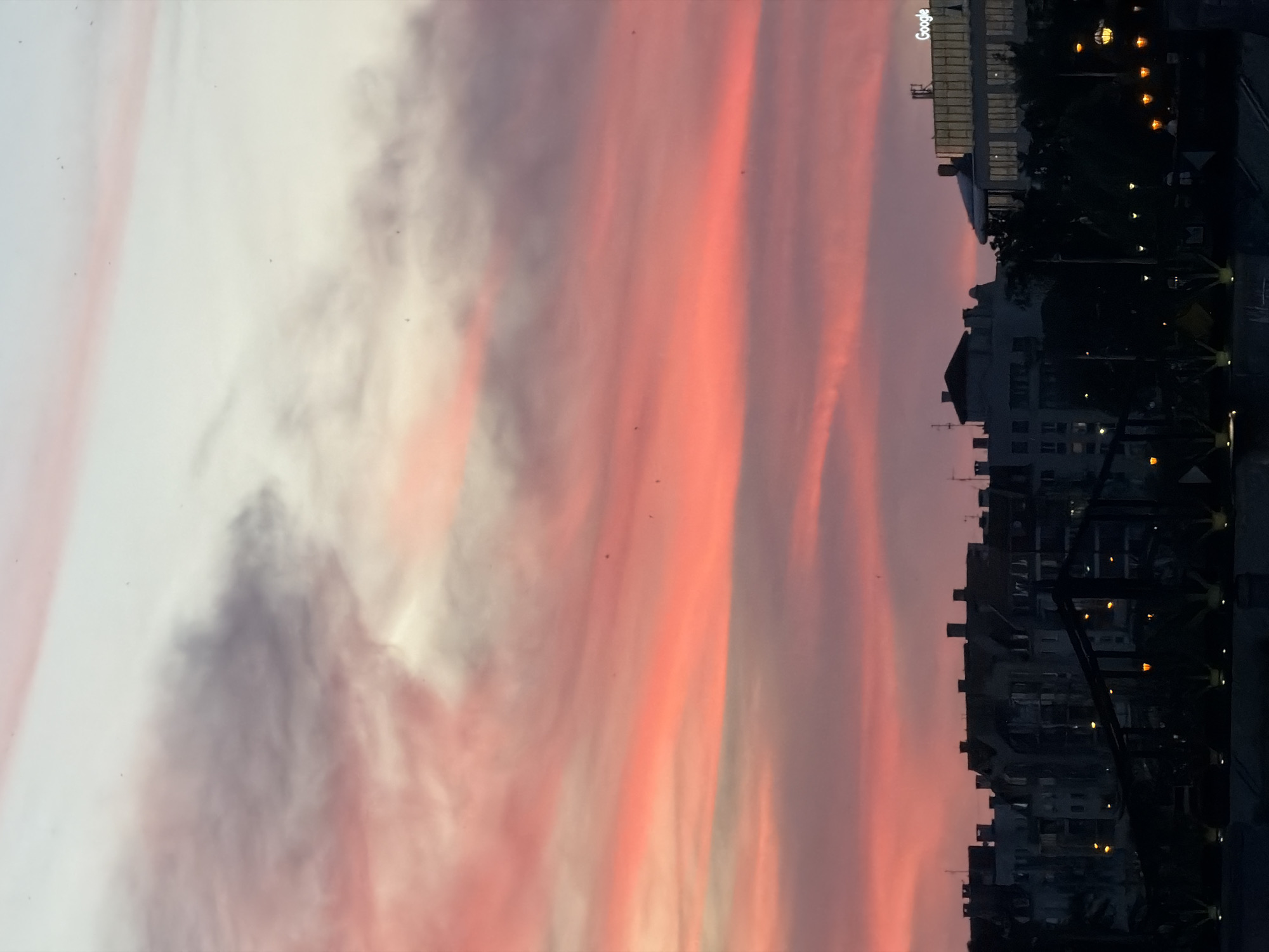 A sunset illuminates the sky over a city skyline in Unknown, Unknown. The sky is filled with wispy clouds painted in hues of pink, orange, and white. Below, the silhouettes of buildings are visible, with a modern glass building on the right displaying the "Google" logo. The lower portion of the image is dark, showing the outlines of trees and lights, suggesting a waterfront or elevated viewpoint.