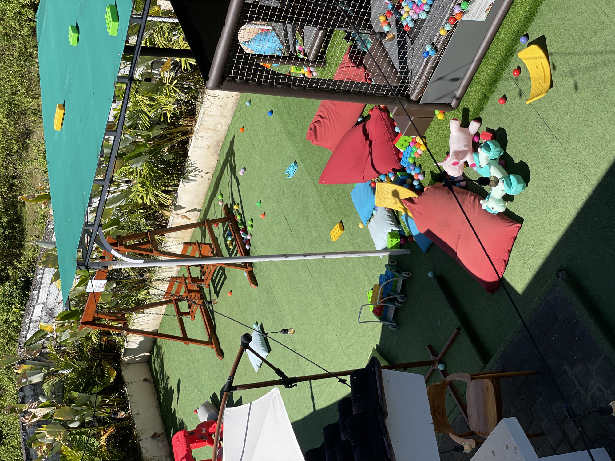 The image depicts a children's play area located in an outdoor setting, presumably in a residential area, city of Unknown, Unknown. The ground is covered with artificial green turf. The scene includes a variety of play equipment and toys, which have been scattered across the area.
On the top right, there is a caged play structure filled with multicolored balls. Several beanbag chairs in shades of red and blue are adjacent to the cage, with a cluster of colorful plastic blocks and toys scattered around them. There are plush toy unicorns laying near the beanbags.
A wooden swing set is located on the left, with green plants and shrubbery in the background, behind the edge of a concrete retaining wall.
Additional toys and blocks are scattered across the green turf. A small stroller with plastic blocks is present, as well as a seesaw-like yellow plastic rocker.
In the foreground, there is a patio umbrella with the supporting structure and chairs beneath it, indicating an adult seating area adjacent to the play area.
Overall, the scene suggests a children's play area with an assortment of toys and equipment, implying active or recent use.