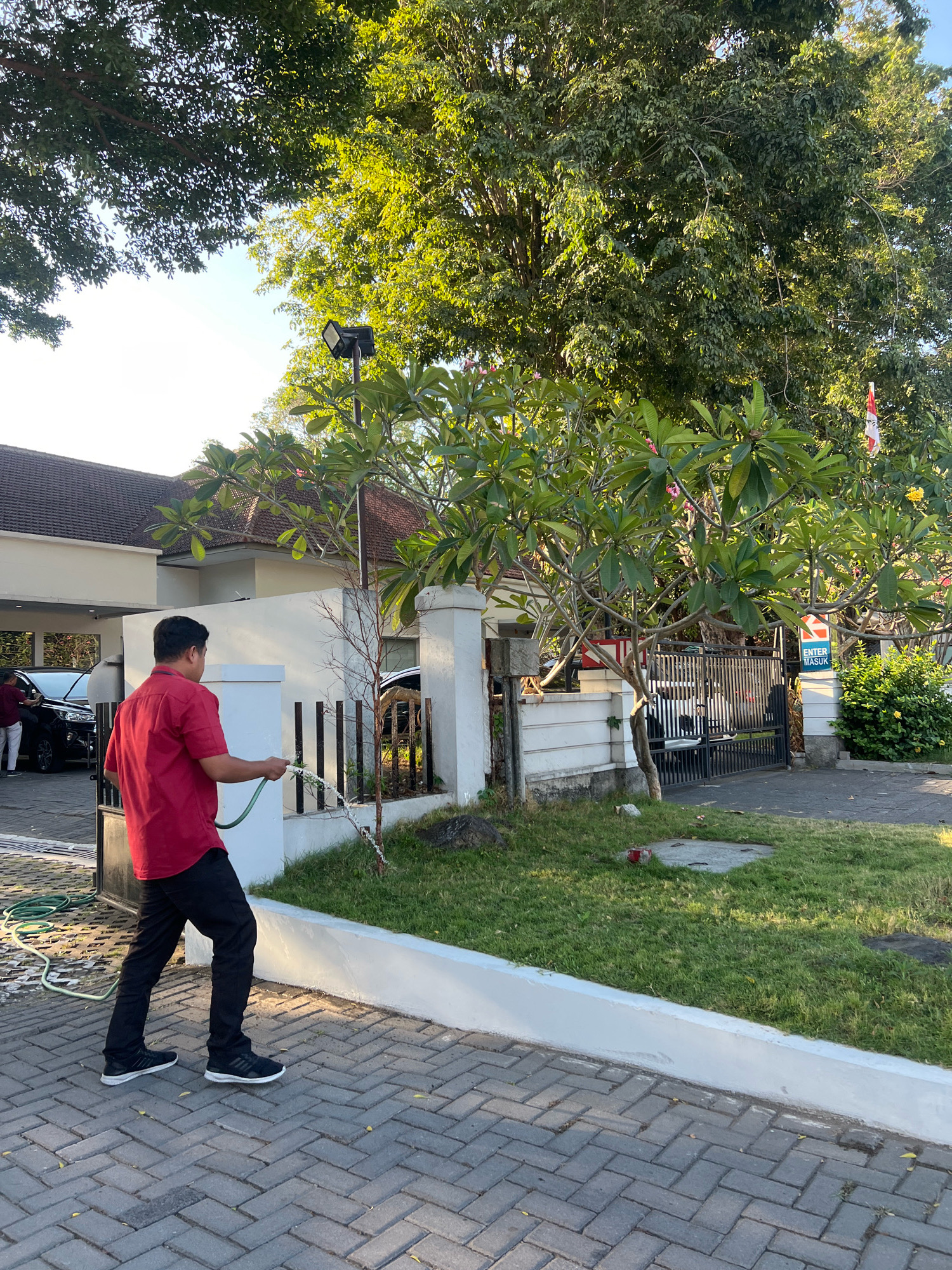 In Depok, Indonesia, a man wearing a red short-sleeved shirt and black pants is watering plants with a green hose. He stands on a brick-paved walkway in front of a white-walled building with a tiled roof. Behind him, a black car is parked under a covered area. The man is facing a fence and a grassy area where he is directing the water. Large trees with green foliage are visible in the background, with sunlight filtering through the leaves. The scene appears to be a residential or commercial property.