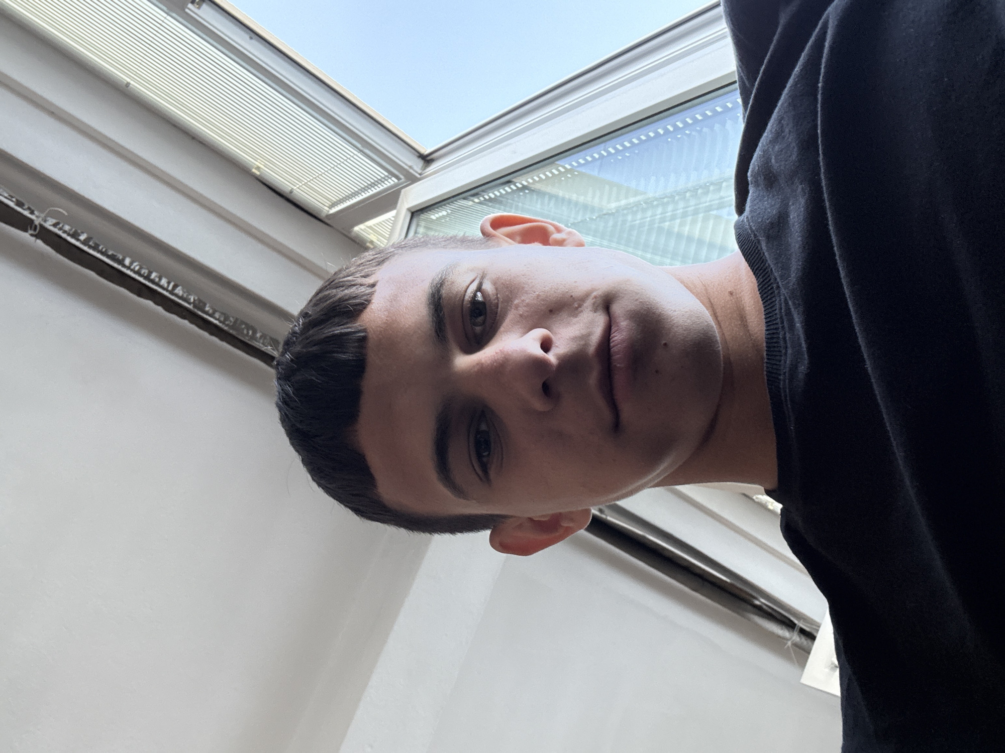 A young man with short, dark hair and fair skin is captured in a close-up selfie. He is wearing a black crew-neck t-shirt. The background suggests an indoor setting with a large window to his right, which offers a view of a bright, clear sky and what appears to be a modern building or structure. A light-colored ceiling is visible above him, with some visible fixtures or structural elements. The angle of the shot is from below, looking up at the subject. The image was taken in Plovdiv, Bulgaria.