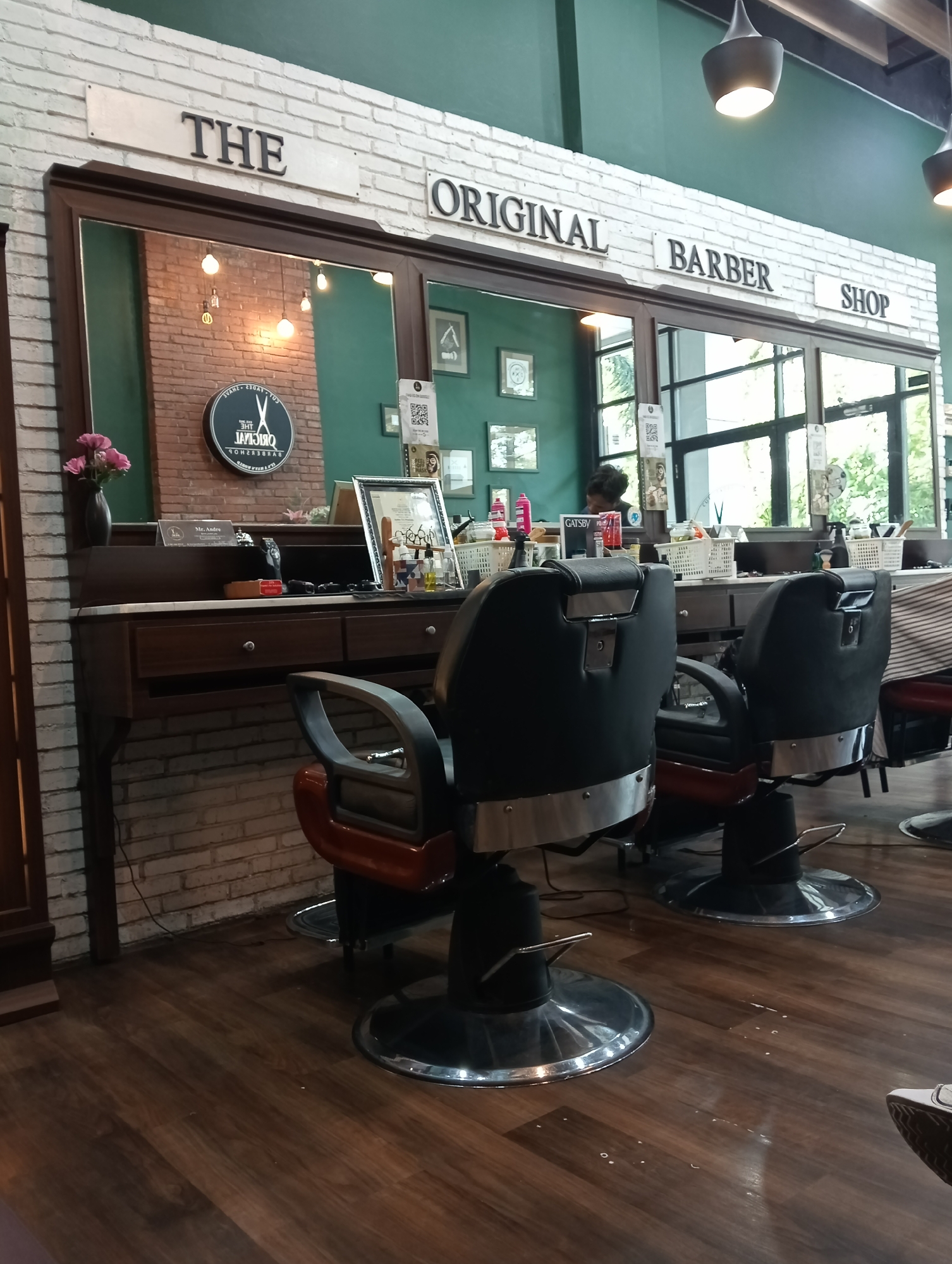 This is a photograph of the interior of a barber shop. The shop appears to be located in Surabaya, Indonesia, as evidenced by a QR code poster on the wall that displays "GATSBY" and a logo with Indonesian text.
The scene is dominated by two barber chairs, both black with chrome accents and adjustable hydraulic bases. The chairs are positioned on a dark wooden floor. Behind the chairs, a long, dark wooden counter or desk is visible, with mirrors above it. The mirrors reflect the interior, including exposed brick walls, hanging light bulbs, and framed pictures on the green wall.
On the counter, various barber tools and products are arranged, including bottles, brushes, and small containers. A vase with pink flowers sits on the left side of the counter. A round, black sign with a logo featuring scissors and text that appears to be "THE ORIGINAL BARBER SHOP" is reflected in the mirror.
The walls are a mix of exposed white brick and a dark green paint. The ceiling has visible wooden beams and industrial-style pendant lights. In the background, through a large window, trees and foliage are visible, suggesting an outdoor area or a bright day.
A person, whose face is not clearly visible, is seen in the background, possibly working at the counter. They appear to be wearing dark clothing. There is also a poster with a QR code and text that mentions "GATSBY," a brand of men's grooming products popular in Indonesia. Another poster features an image of a man with text that is difficult to read but likely related to grooming services.
The overall impression is of a modern, yet somewhat rustic, barber shop with a focus on vintage aesthetics, indicated by the barber chairs and the brickwork. The lighting is warm, with both natural light from the window and artificial light from the pendant lamps. The atmosphere appears clean and organized.