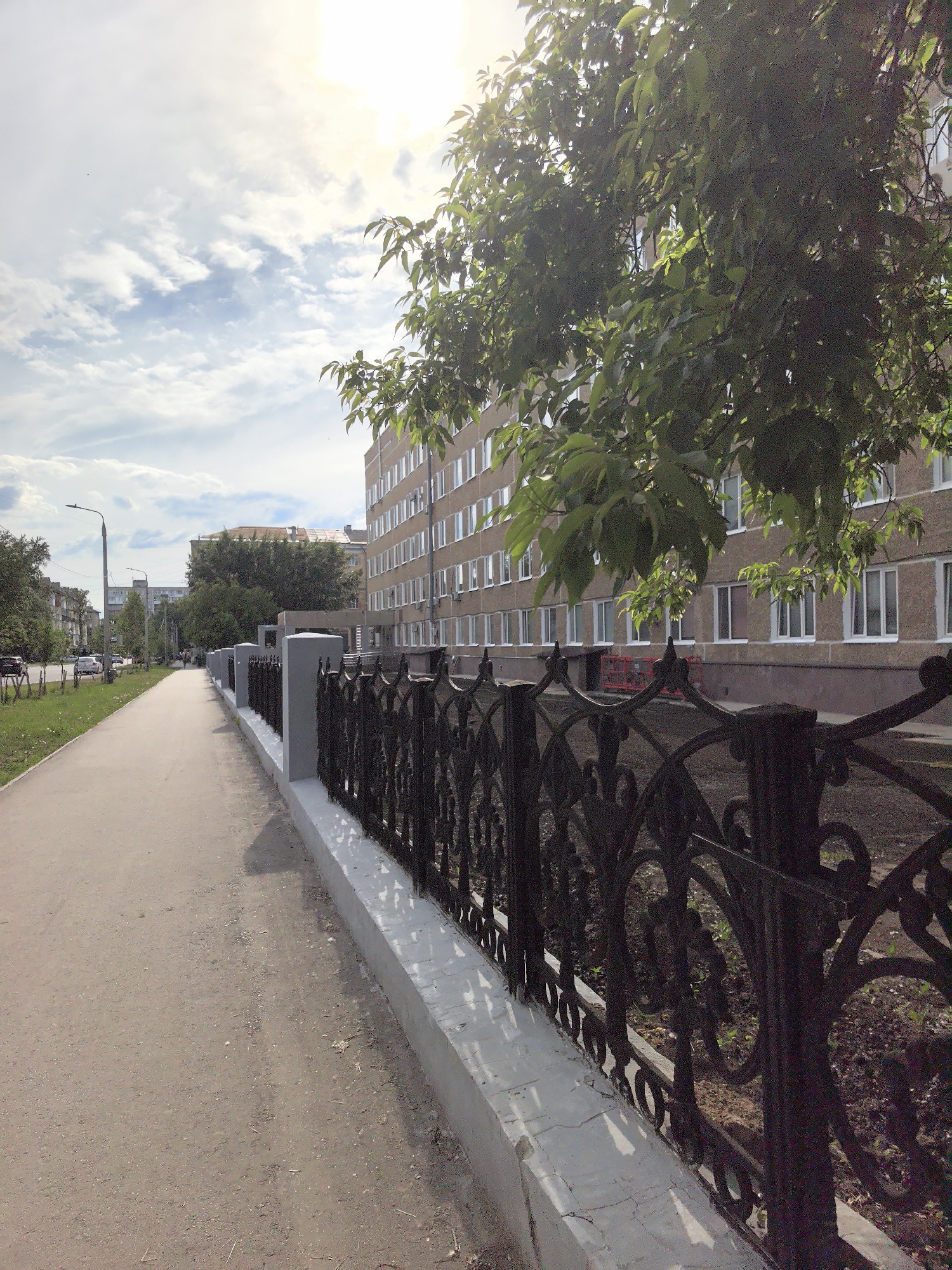 The image depicts a bright, daytime outdoor scene, likely in an urban or suburban environment, characterized by a long pedestrian path running alongside a street and buildings.
In the foreground and extending into the midground on the left, a wide, light brown dirt or gravel sidewalk stretches into the distance. To its immediate right, a dark, elaborately designed metal fence with intricate, swirling scrollwork patterns stands on top of a low concrete wall, which is painted white on its upper surface and grey below. The fence casts sharp shadows onto the wall and the ground. Behind the fence, the ground appears to be an unkempt area of dark earth with some sparse green plant shoots.
On the right side of the frame, a multi-story building, likely an apartment block or office building, is partially visible. It has a light brown or beige facade with numerous evenly spaced, rectangular windows with white frames. A large tree with vibrant green foliage largely obscures the building, particularly in the upper right. Bright sunlight filters through the tree's leaves, causing some lens flare and illuminating the foliage.
The sky in the upper left is bright blue with scattered, fluffy white clouds. To the left of the sidewalk, a narrow strip of grass separates the path from a street where several cars are parked in the distance. A tall street lamp is also visible along the street. Other buildings can be seen in the background, lining the street on both sides.
No people or specific activities are distinctly visible in the scene. The time of day appears to be sunny, possibly morning or late afternoon, given the angle of the light and shadows. There is no visible text or signage in the image. The location context is unknown.